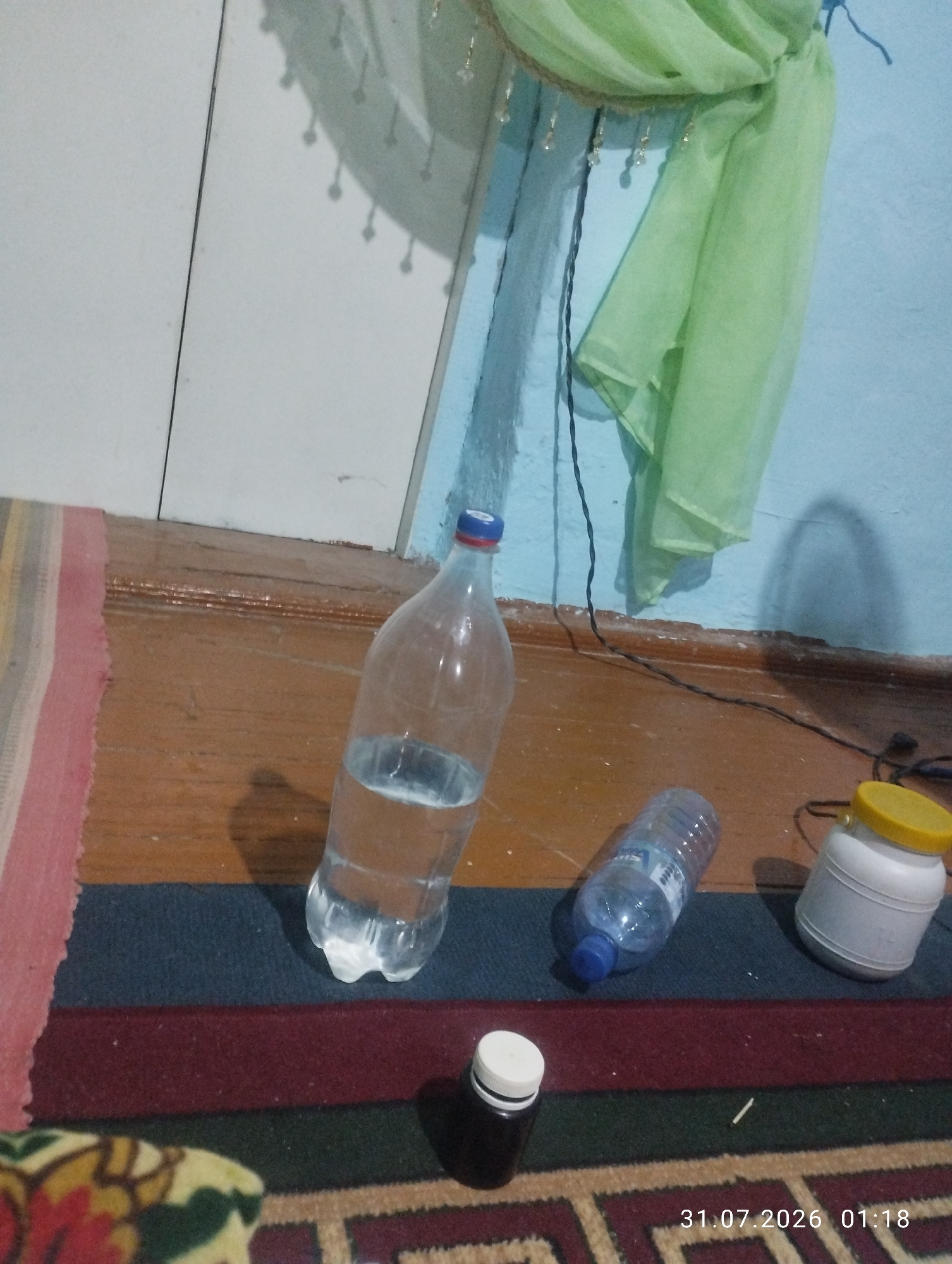 The image shows a scene indoors, likely a room. On a wooden floor, a large clear plastic bottle filled with water is placed. To its right, a smaller clear plastic bottle with a blue cap is visible, and further to the right, a white plastic container with a yellow lid is present. In the foreground, a small dark bottle with a white cap sits on a carpet. In the background, a light blue wall meets a wooden wall panel. A piece of light green fabric is draped on the wall. A date and time stamp, "31.07.2026 01:18", is visible in the bottom right corner. The location is identified as Quyosh, Uzbekistan.