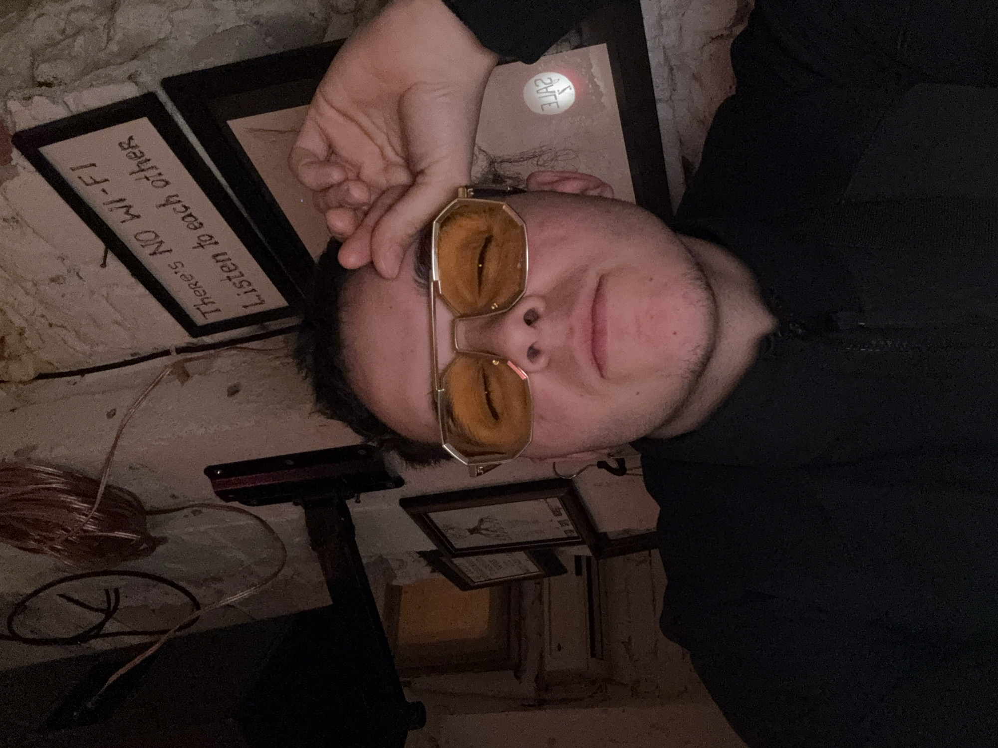 The image, taken from an overhead perspective, shows a person lying on their back, looking up towards the camera. The individual, appearing male, has dark hair and light facial stubble, and is wearing distinctive octagonal-shaped eyeglasses with yellow-orange tinted lenses. One hand rests casually near their head, partially obscuring their ear. They are dressed in a dark, possibly black, long-sleeved top or jacket with a visible zipper.
The person is situated on a textured, light-colored surface, resembling a rough concrete or stone floor. The lighting is dim, suggesting an indoor setting, possibly in the evening or a space with subdued illumination, characteristic of a cozy establishment in Sofia, Bulgaria.
Several objects are visible around the subject. Near their head, two framed items are placed on the surface. One prominently displays the text, "There's NO WI-FI / Listen to each other," printed upside down relative to the person's orientation. Another frame shows a faint, mirrored word that appears to be "SALE." Further into the background, additional framed pictures or signs are scattered on the surface. A coil of reddish-brown wire and a dark, metallic object are also discernible.
The scene suggests a relaxed atmosphere in a unique indoor environment, possibly a cafe, bar, or a distinctive room, emphasizing offline social interaction. The person's expression is neutral to slightly content, with a hint of a smile.