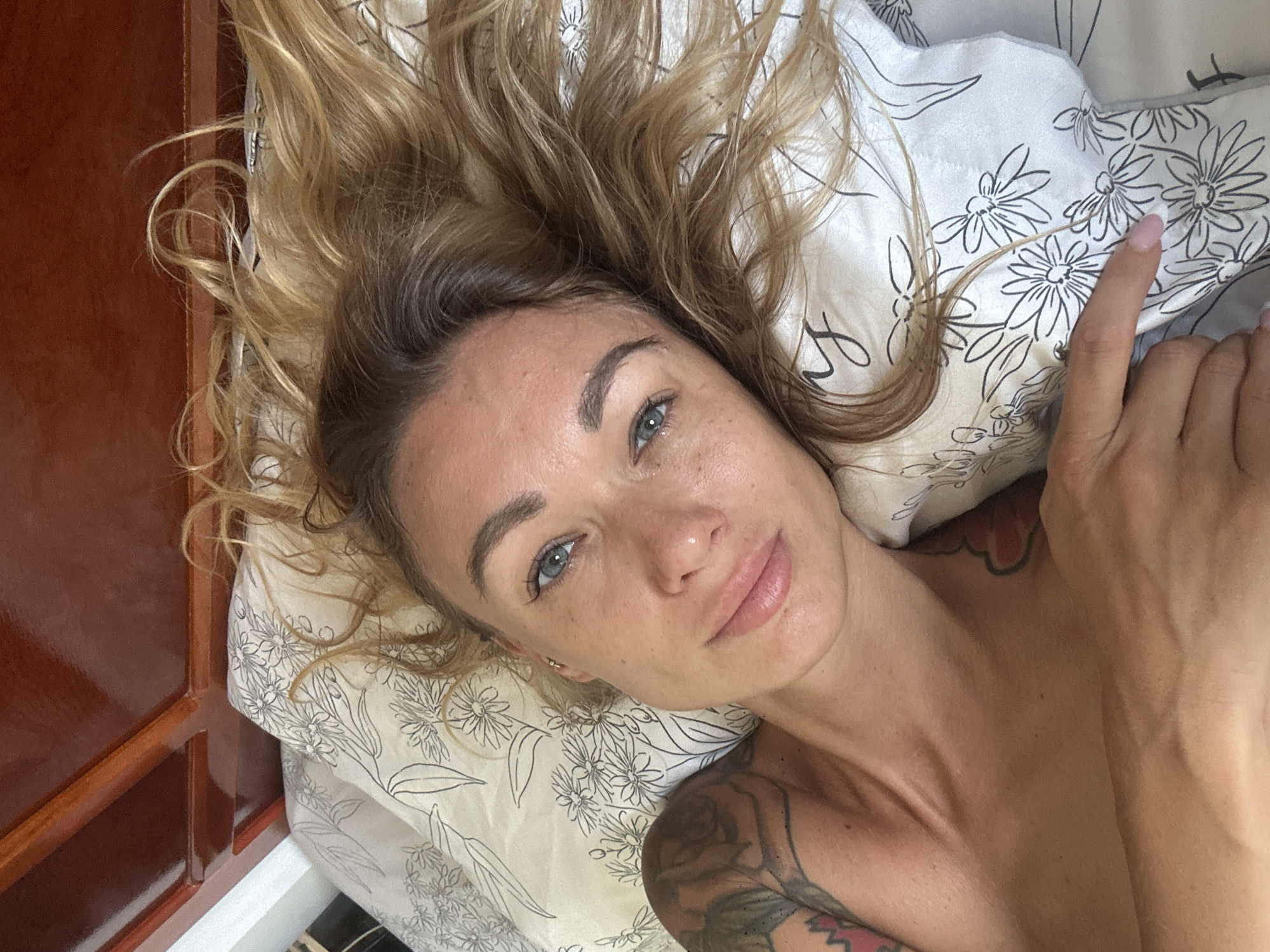 A woman with blonde hair and blue eyes lies on a bed with floral patterned sheets. Her left arm is visible, showing tattoos. Her head is resting on a pillow. The scene appears to be indoors. There is no indication of the city of Mũi Né, Vietnam.