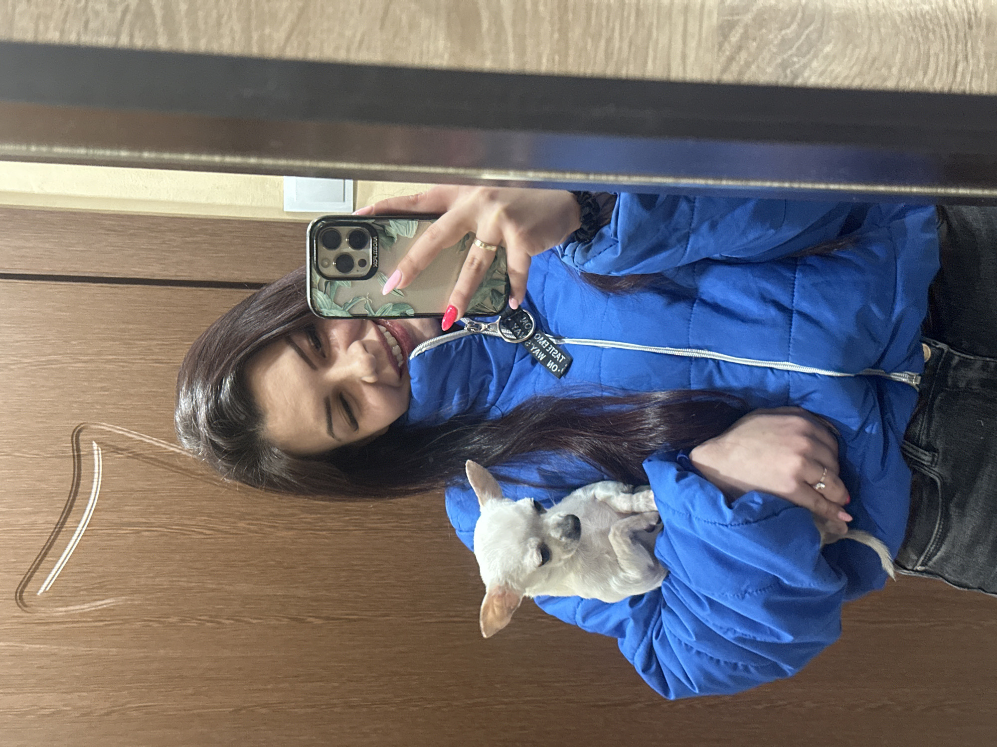 A young woman is taking a selfie in a mirror. She is wearing a blue jacket and holding a small, white dog. The background is a wooden door. The location is likely indoors, but not definitively Sliven, Bulgaria.