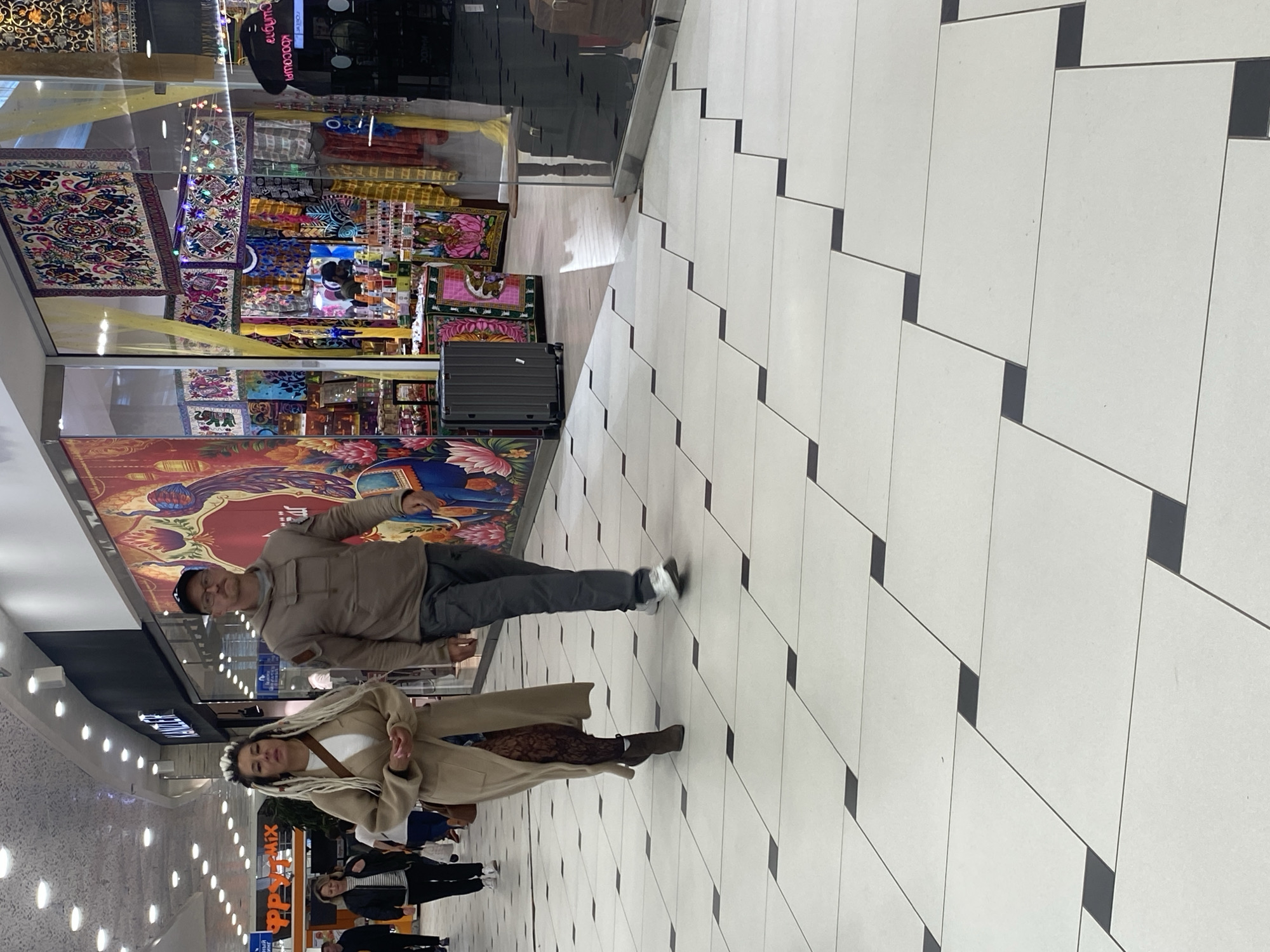 The image captures an interior view of what appears to be a shopping mall or large retail space, possibly within the city of Yekaterinburg, Russia. The foreground features a floor tiled with white, rectangular tiles arranged in a staggered pattern with black accents. Two individuals are the main subjects: a man wearing a tan jacket, dark pants, and a black hat, and a woman with long, light-colored hair, wearing a light tan coat over a white shirt and a brown patterned skirt. They appear to be walking. Behind them, there are other people walking in the same direction, suggesting a flow of pedestrian traffic. To the left, there are shops and signage, including the word "Парк" in orange lettering, indicative of the retail setting. Behind the man, a large display area showcases colorful textiles and decorative items, potentially for sale. A black suitcase stands near the display. The environment suggests a bustling commercial area with a variety of retail offerings.