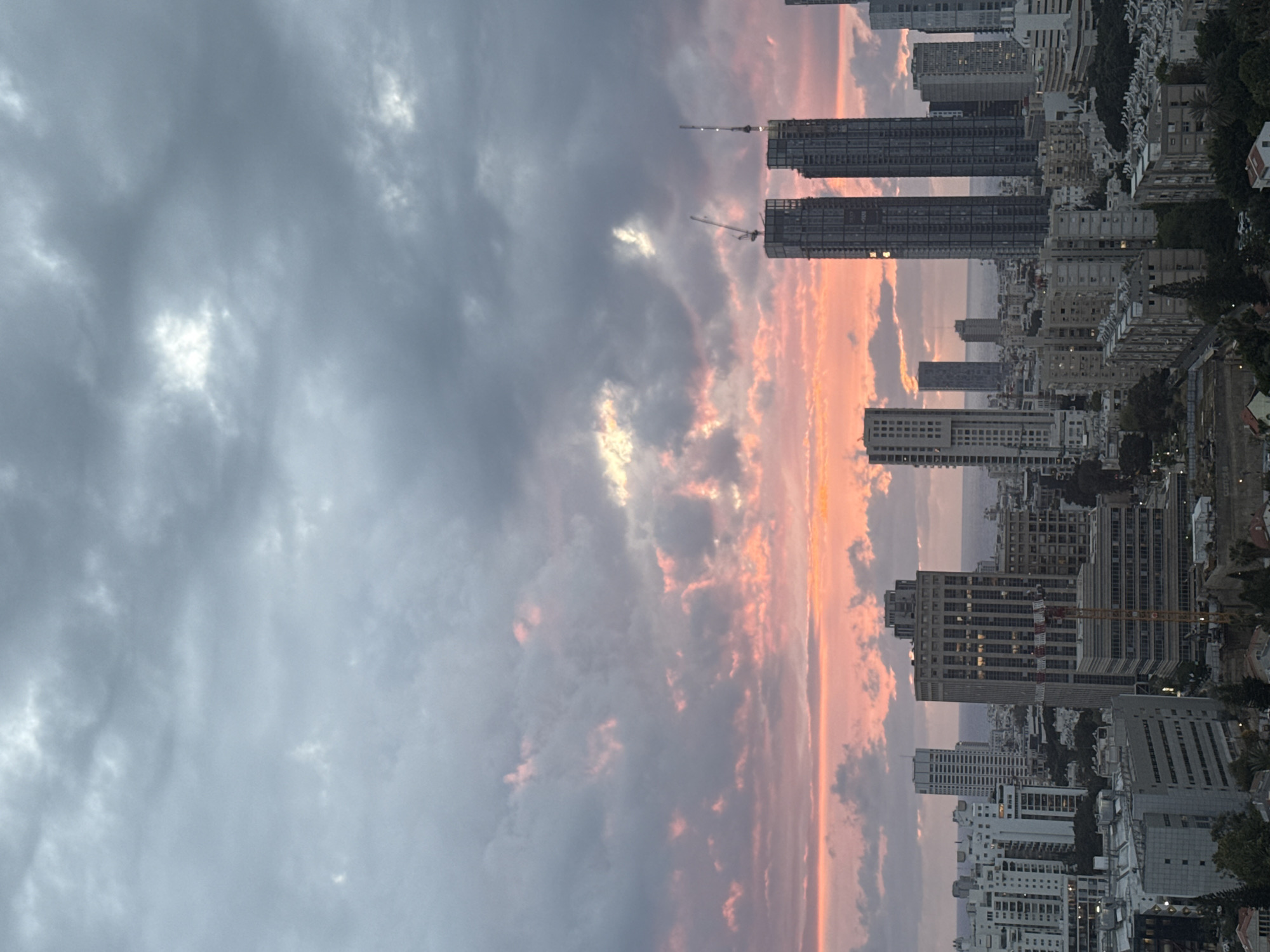 The image depicts a cityscape at dusk, with a prominent skyline and a dynamic cloudscape. Tall buildings dominate the view, showcasing the urban architecture. Construction cranes can be seen near the buildings, indicating ongoing development. A sunset casts a warm, pinkish-orange hue across the clouds, contrasting with the darker, more subdued tones of the city and sky. The location is likely Tel-Aviv, Israel, given the architectural style and coastal setting.