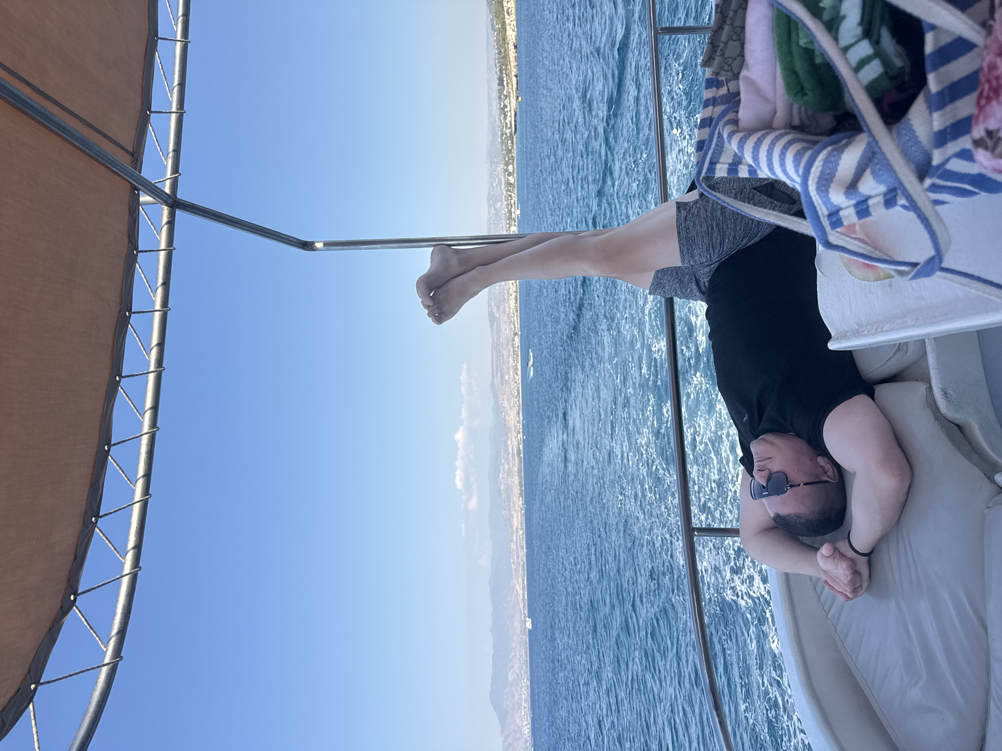 A man is lying on his back on a boat, with his legs raised and extended straight up in the air. He is wearing a black t-shirt and grey shorts. His arms are crossed behind his head. The boat is on the sea, with a clear blue sky and distant mountains visible in the background. The location appears to be near Neo Chorio, Cyprus, given the coastline and mountainous terrain. There is a striped bag with towels and clothing next to him.