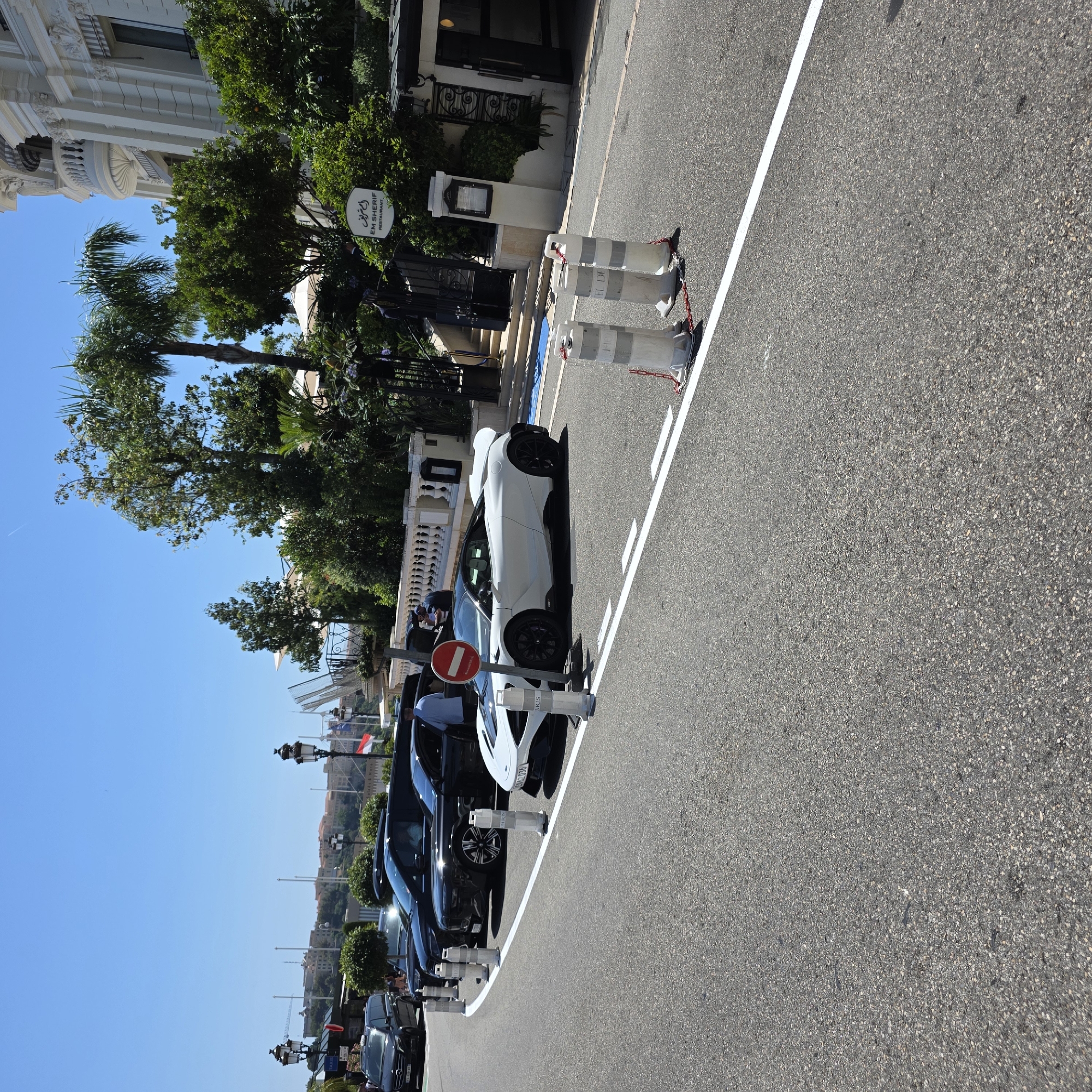 In Monaco, Monaco, a white McLaren sports car is parked on a road. The car has a black roof and black accents. It is parked behind a black luxury SUV and a black sedan. A person in a blue shirt is standing near the driver's side door of the black sedan, and another person is standing behind the white McLaren. A red and white circular sign is mounted on a pole in front of the McLaren. The road is lined with white bollards and a white line. In the background, a white building with ornate architecture is visible, along with trees and a blue sky.