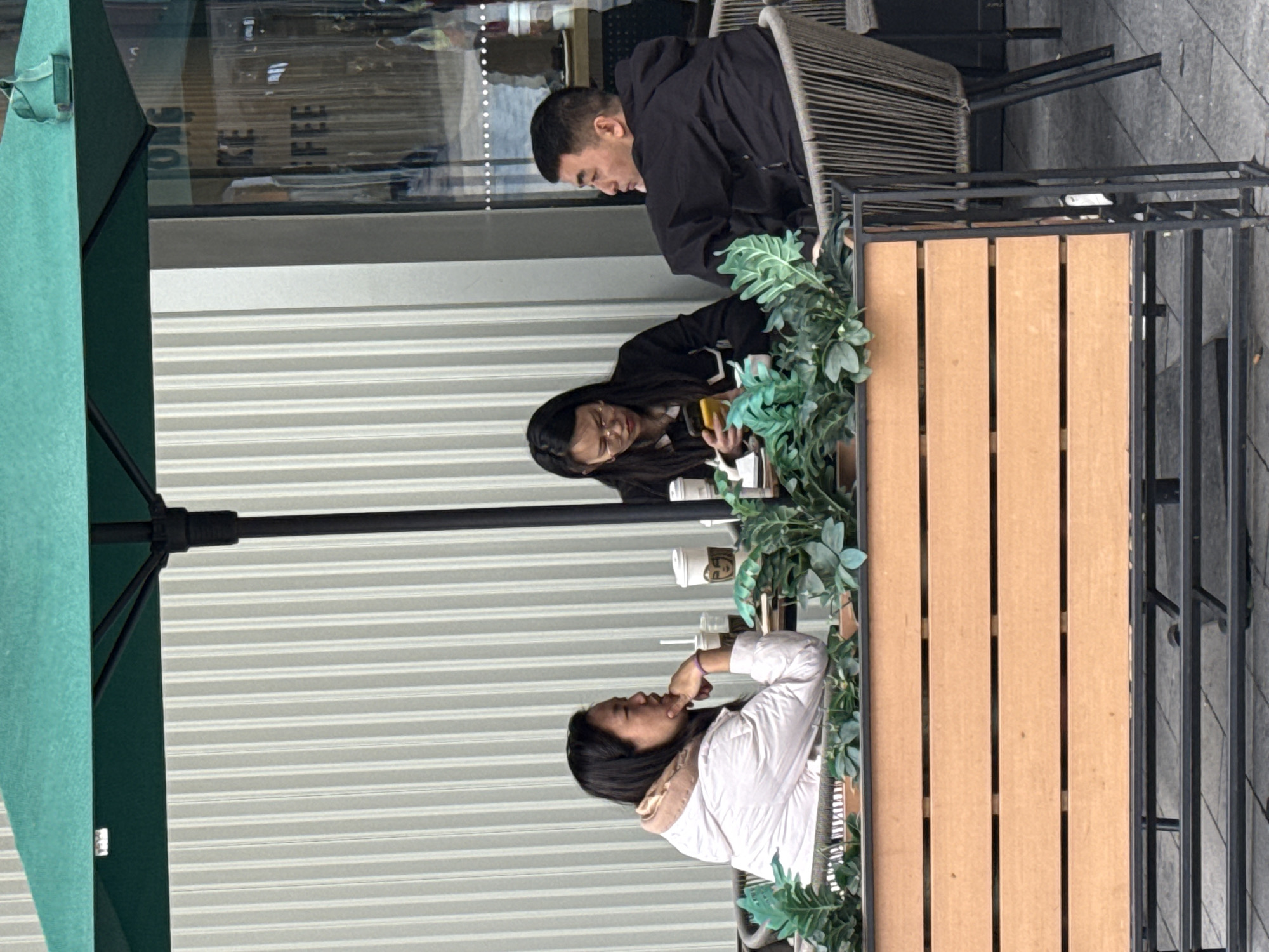 Three people are seated at a table on an outdoor patio, under a green umbrella. The patio is outside of what appears to be a cafe in the Yuzhong District, China. The table is rectangular, made of wooden planks surrounded by a black metal frame, decorated with faux greenery. A man in a black hooded jacket is seated furthest from the camera, looking down at the table. A woman in a black jacket and glasses is sitting in the middle, looking at her phone. A woman in a white jacket is seated closest to the camera, looking upwards and touching her mouth with her hand. There are several paper cups on the table, indicating they may have purchased drinks from the cafe.