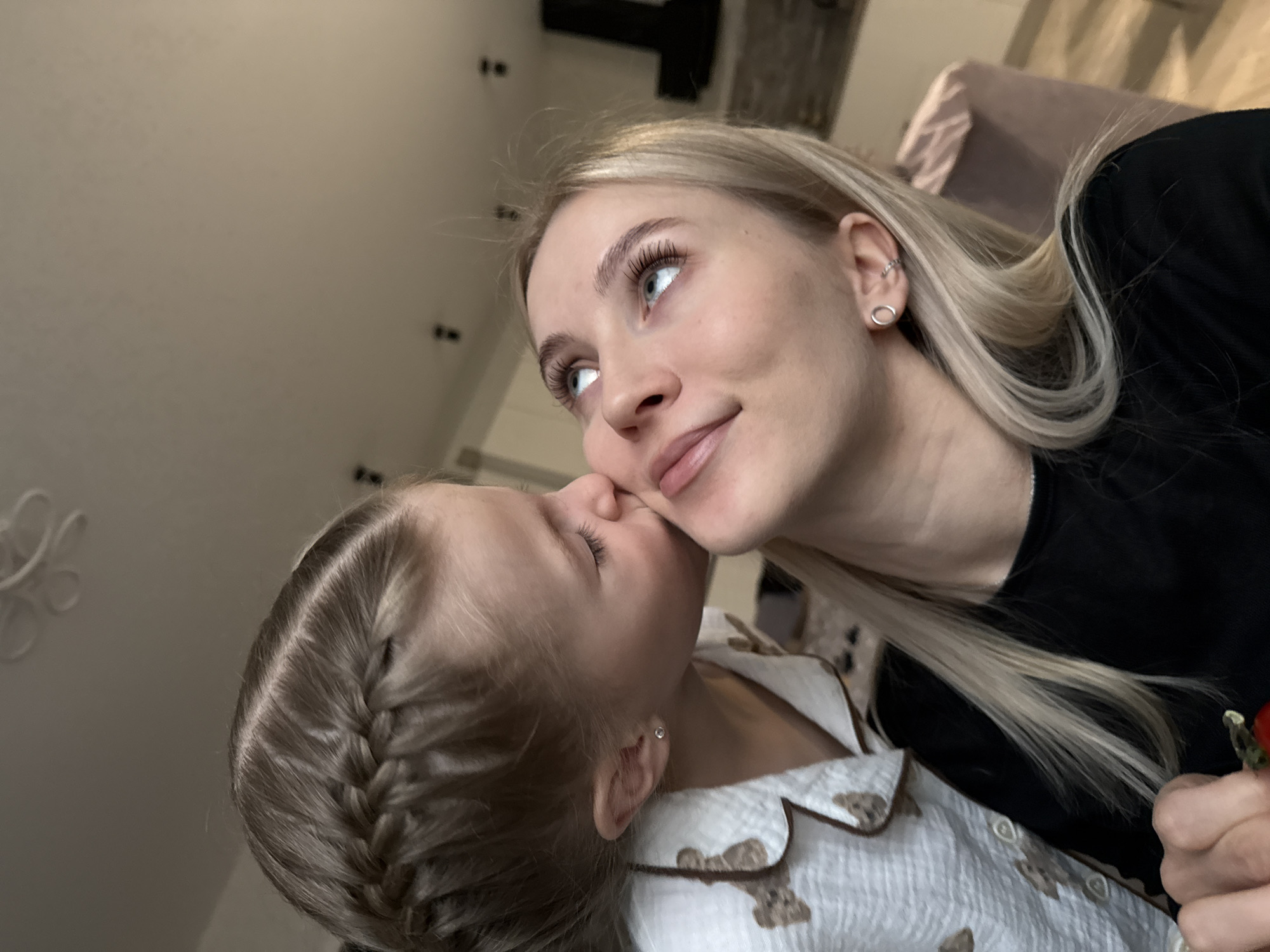 A woman with blonde hair and a black shirt is being kissed on the cheek by a child wearing a light-colored shirt with a bear pattern. The woman is looking upward to the left. The location cannot be determined from the image.