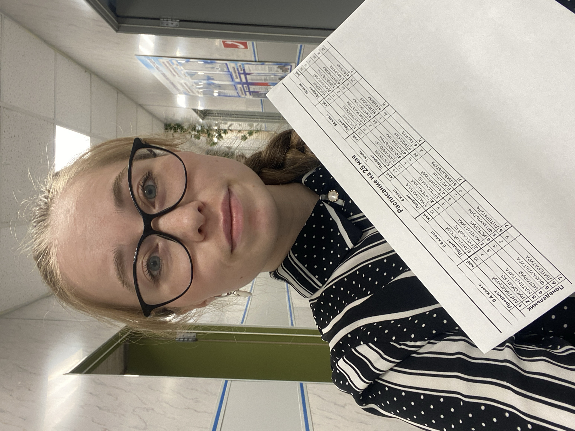 A woman is holding a sheet of paper with a school schedule on it. The paper reads "Schedule for May 25". The table contains the subjects that will be taught on that day for classes 5A, 5B, 6th, 7th, 8A, and 8B. Subjects listed include English, Russian language, Math, History, PE, Literature, Physics, and Biology. The location is likely a school hallway, with bulletin boards and tile walls visible in the background. The location is possibly in Saint Petersburg, Russia, as the schedule is written in Russian.