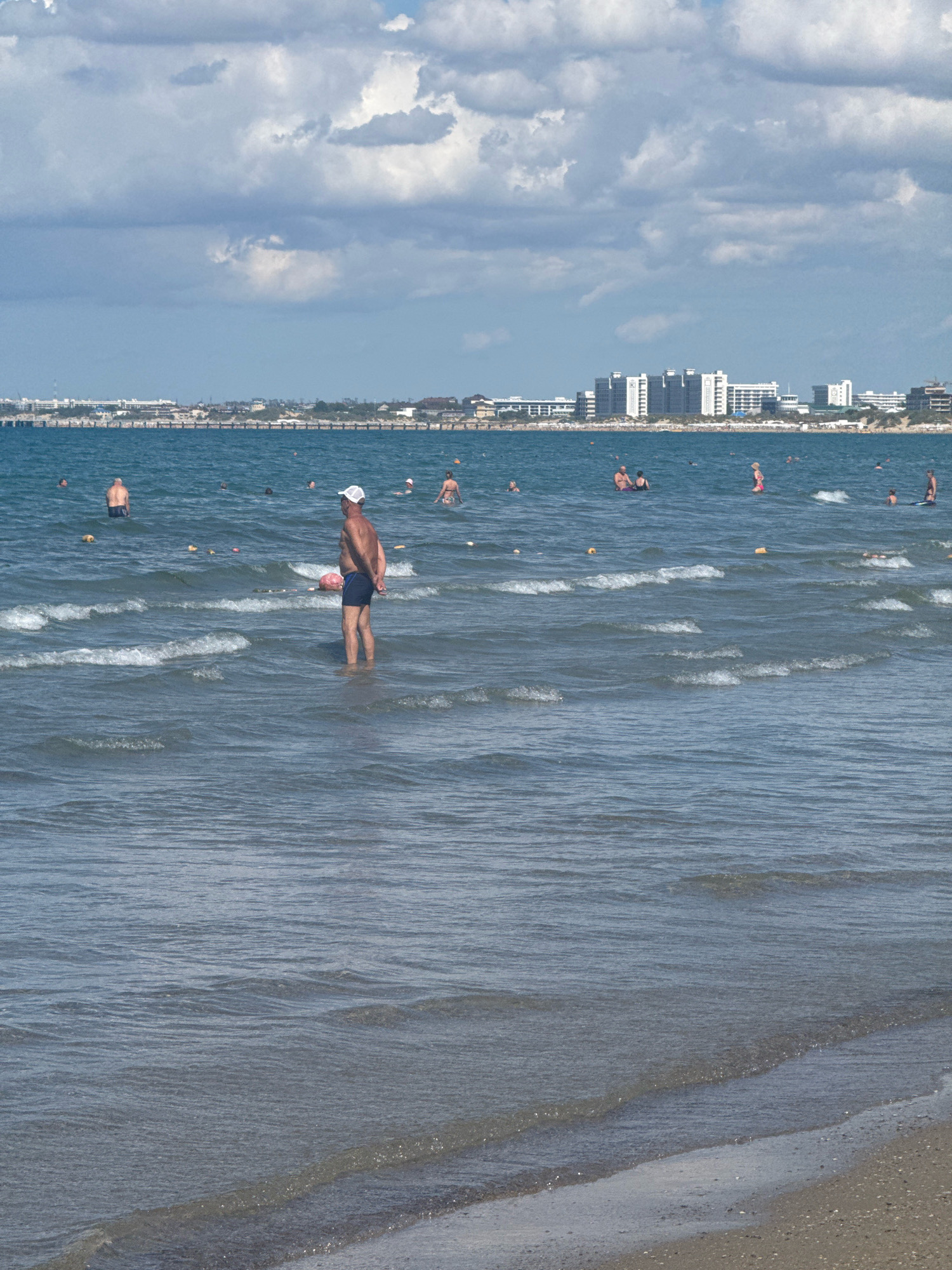 This is a medium-distance, eye-level shot of a beach in Anapa, Russia, on a partly cloudy day. The foreground is dominated by the sea, with gentle waves lapping at the shore. Several people are in the water, some wading and others swimming further out. The middle ground shows a line of buoys and a large pink inflatable object. In the background, a coastline is visible, lined with numerous multi-story buildings, suggesting an urban area. The sky is filled with large, fluffy white and grey clouds.