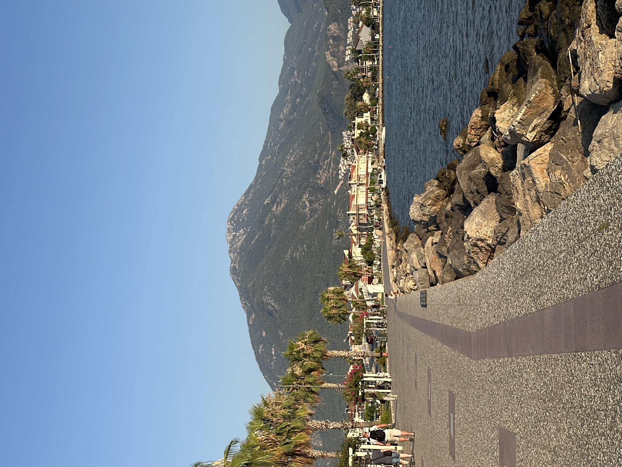 This media file depicts a coastal promenade with a mountainous backdrop, likely in Fethiye, Turkey. The scene is characterized by a clear blue sky, suggesting fair weather and possibly a warm time of day, given the long shadows cast by the palm trees lining the promenade.
The primary subjects are the natural elements of the landscape: a large, green, and sparsely vegetated mountain dominates the background, meeting the deep blue sea on the right. The shoreline is fortified with large, rough-hewn rocks, forming a natural breakwater. To the left of the rocks, a paved promenade stretches into the distance. This promenade is lined with palm trees, some with full fronds and others showing more weathered trunks, hinting at a tropical or Mediterranean climate.
Beyond the palm trees, a strip of buildings can be seen, suggesting a town or resort area. These buildings appear to be of a few stories high, with light-colored facades and balconies, characteristic of coastal architecture. Small boats or vessels are visible in the water near the shore.
Several people are present on the promenade, engaged in leisurely activities. Some are walking, while others appear to be sitting or standing, possibly enjoying the view or waiting for something. Their attire is casual, consistent with a warm climate. The overall mood conveyed is one of relaxation and vacation. The time of day appears to be late afternoon or early evening, as indicated by the low angle of the sun and the length of the shadows. No visible text is present in the image.