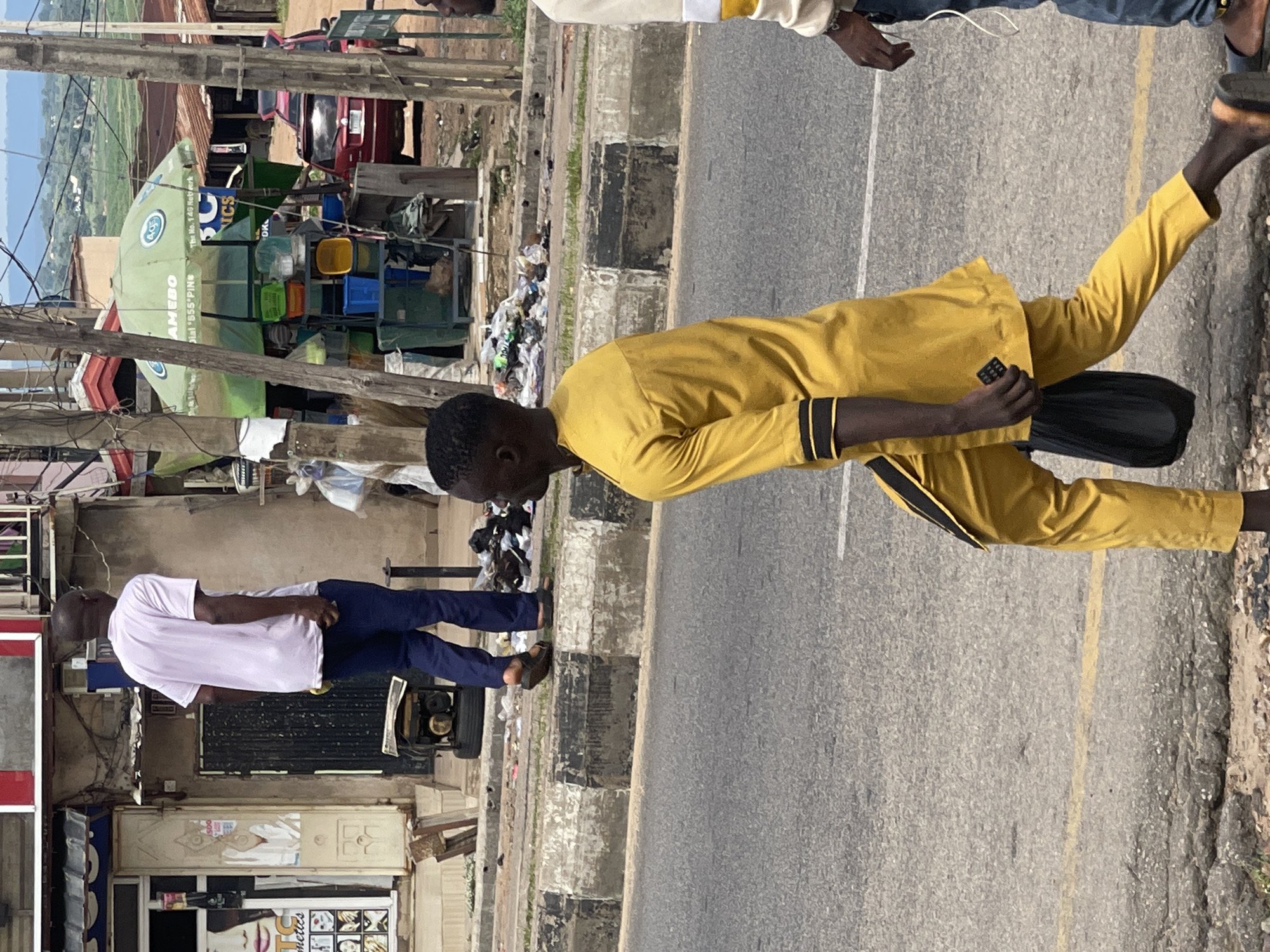 The image shows a street scene with two men. The man in the foreground is wearing a bright yellow outfit and is walking on a paved road. He is holding a dark object in his right hand. The second man is in the background, to the left, wearing a white t-shirt and dark pants. He appears to be standing near some shops.
The setting is an outdoor urban environment with buildings and shops lining the street. There are also some vehicles visible in the background, including a red car. The road has yellow and white markings, suggesting it is a street or highway. There is debris and trash scattered along the side of the road and near the buildings, indicating a potentially unkempt area. A large green umbrella with text on it is prominent in the background, possibly associated with a vendor or business.
The lighting suggests it is daytime, possibly mid-morning or afternoon, with bright sunlight casting shadows. The weather appears to be clear. There are no strong emotional cues visible on the faces of the men.
Visible text includes: "AMCHO" on the green umbrella, and "TELE. NO. 149 NETWORK". There is also text on signage in the background related to "COSMETICS", accompanied by images of faces.