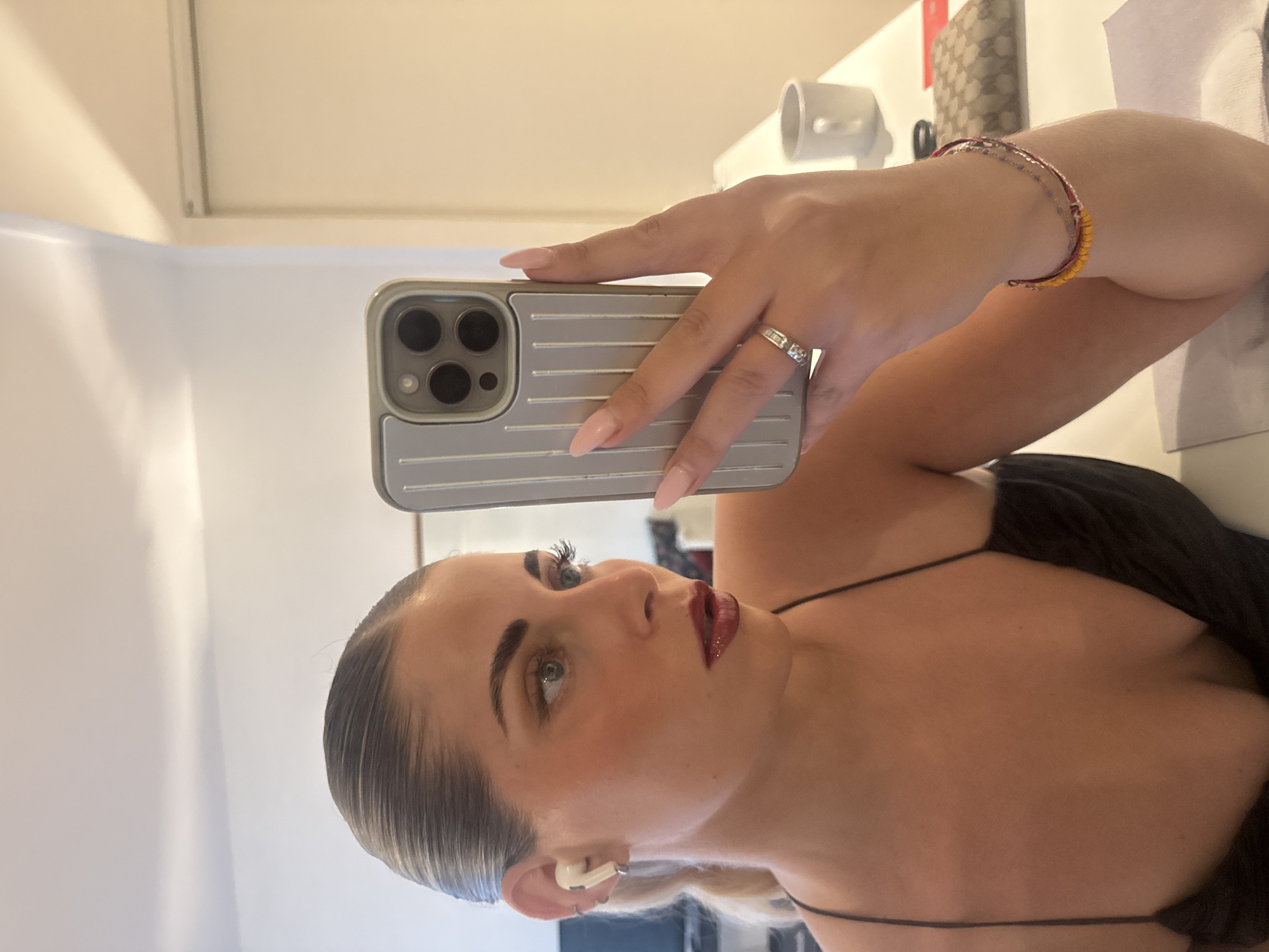 A woman with blonde hair pulled back in a ponytail takes a selfie in a room. She is wearing a black slip dress, white AirPods, and several bracelets. She has on makeup, including lipstick and eyeshadow. Her right hand is holding an iPhone with a silver case, and she is wearing a ring on her left ring finger. In the background, a white mug and a designer wallet can be seen on a white desk. The scene appears to be indoors, possibly a hotel room, with plain walls and a ceiling light. There is no indication of the location being Paris, France.
