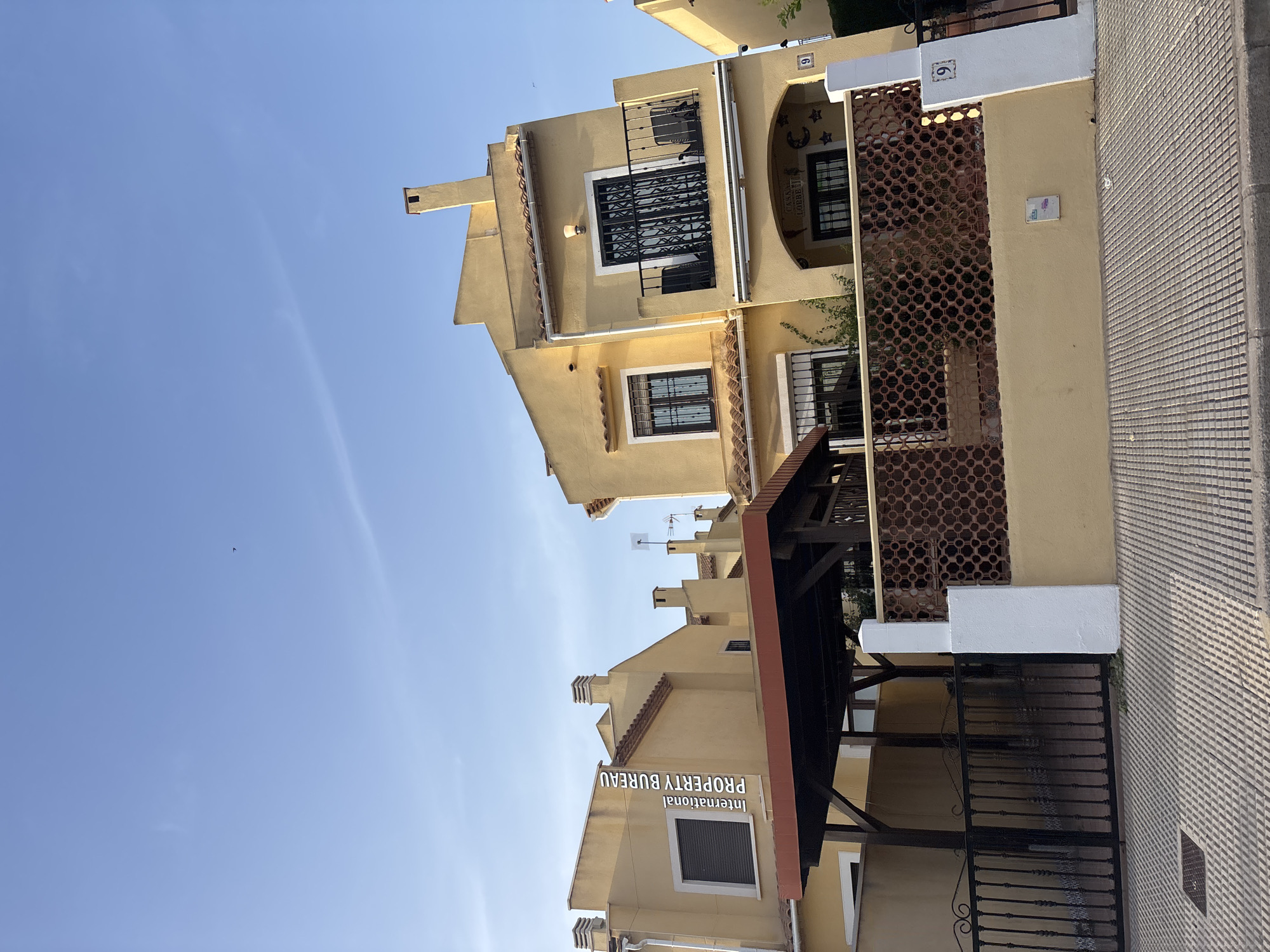 The image shows a street view of a row of attached houses in Orihuela, Spain. The houses are painted a light yellow color and have red tile roofs. Each house has multiple stories, windows with dark shutters, and chimneys. One house has a balcony with black metal railings. The houses are bordered by a decorative brown metal fence and a low yellow wall. A black metal gate is visible in the foreground, leading to a covered parking area. The street in front of the houses is paved with light-colored tiles. A white post with a house number "9" is visible to the right of the image. The sky is clear and blue, with a few wisps of clouds and a single bird in flight. A watermark reading "International PROPERTY BUREAU" is overlaid vertically on the left side of the image. The overall impression is a residential street scene on a sunny day.