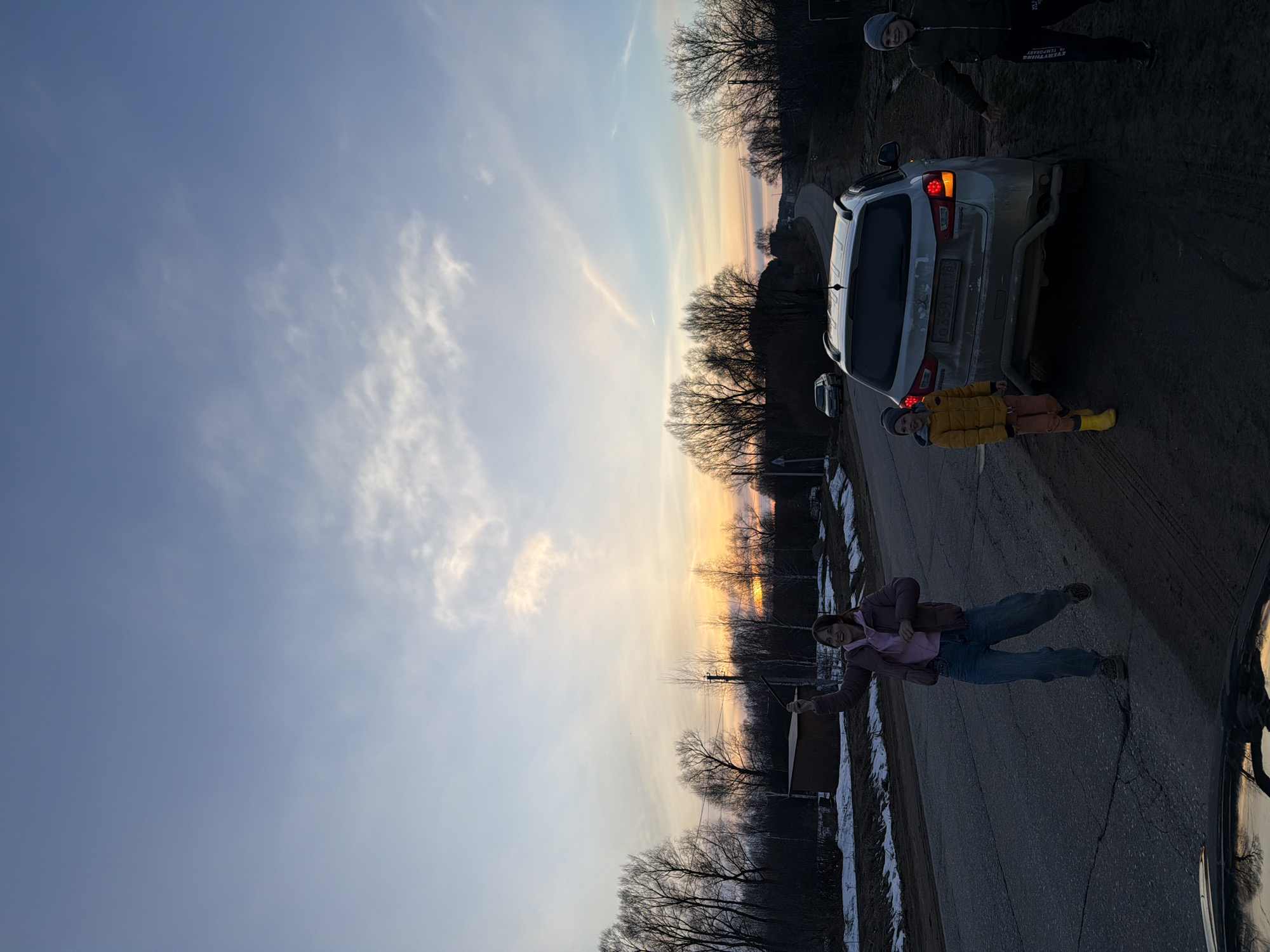 The image shows a road in Дулесово, Russia, at sunset. A silver car is parked on the side of the road. Three people are present: a person in a pink jacket and jeans stands on the road, a child in a yellow jacket and orange pants stands near the rear of the car, and another person in a dark jacket stands further away from the car. Trees are visible along the roadside, silhouetted against the colorful sky. Patches of snow are on the side of the road.