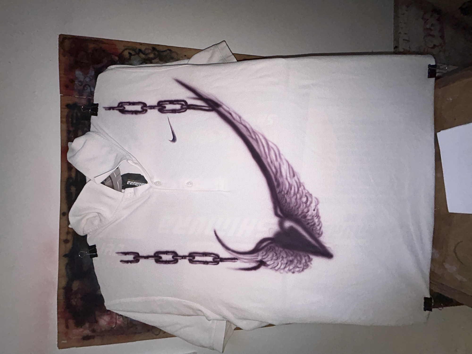 A white polo shirt with a purple design resembling a wing and chain is clipped to a paint-splattered wooden board. The background includes parts of a white wall and a cardboard box. No people or location are identifiable. There are no other elements of Buenos Aires visible in the image.
