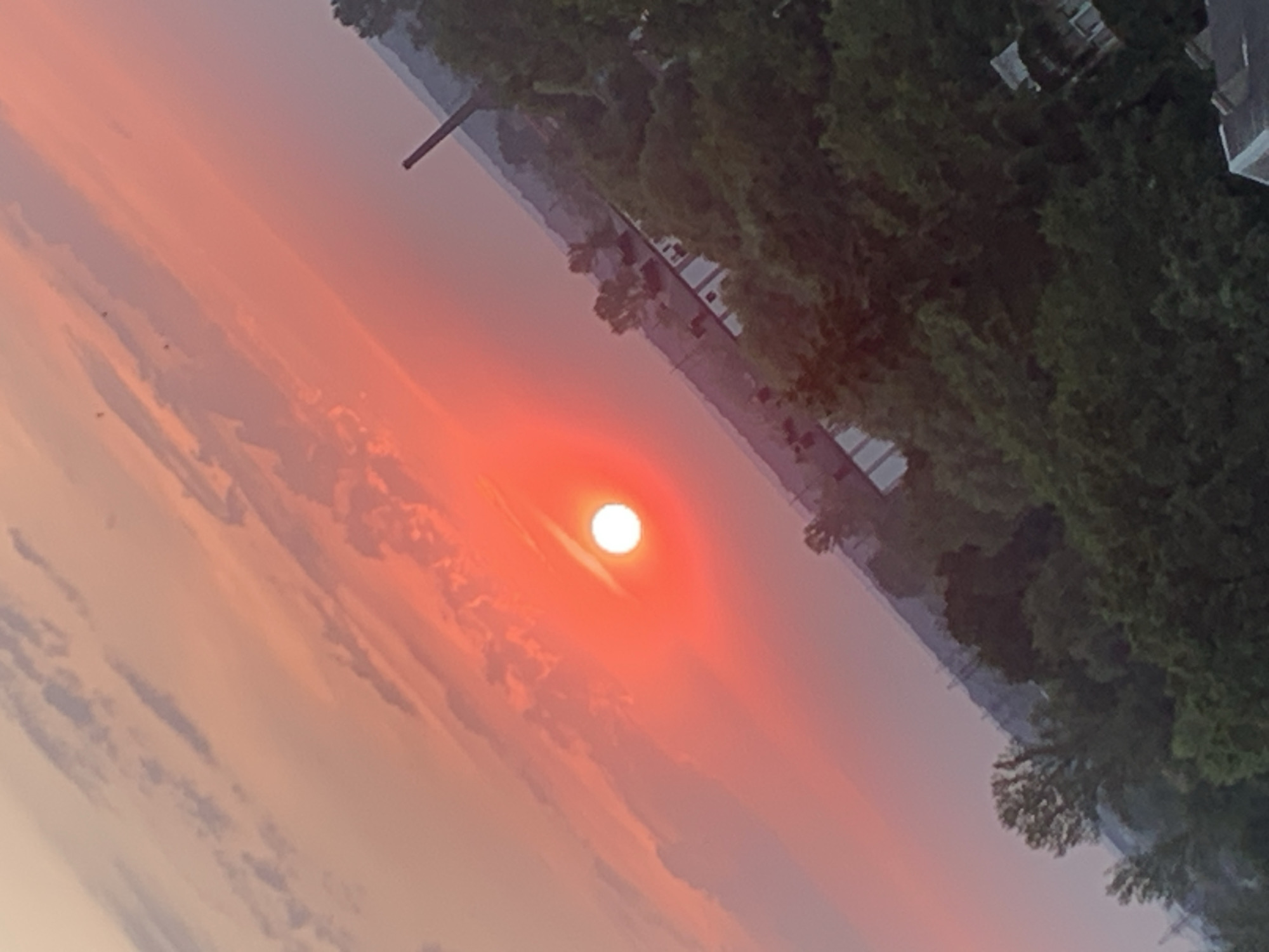 The image depicts a vibrant sunset over a city, with the sun appearing as a bright, orange-red orb partially obscured by wispy clouds. The sky transitions from a deep orange at the horizon to lighter shades of pink and pale yellow higher up. In the foreground, a dense canopy of green trees is visible, suggesting a natural or park-like area within the city. Several buildings with white walls and dark roofs are interspersed among the trees, indicating a residential or mixed-use urban environment. A tall, dark smokestack or industrial chimney rises in the background, partially silhouetted against the colorful sky. The scene is captured at an angle, giving a dynamic perspective of the landscape. There are no people or animals visible in the frame. The overall impression is one of a picturesque sunset in an urban setting, possibly during the late afternoon or early evening.