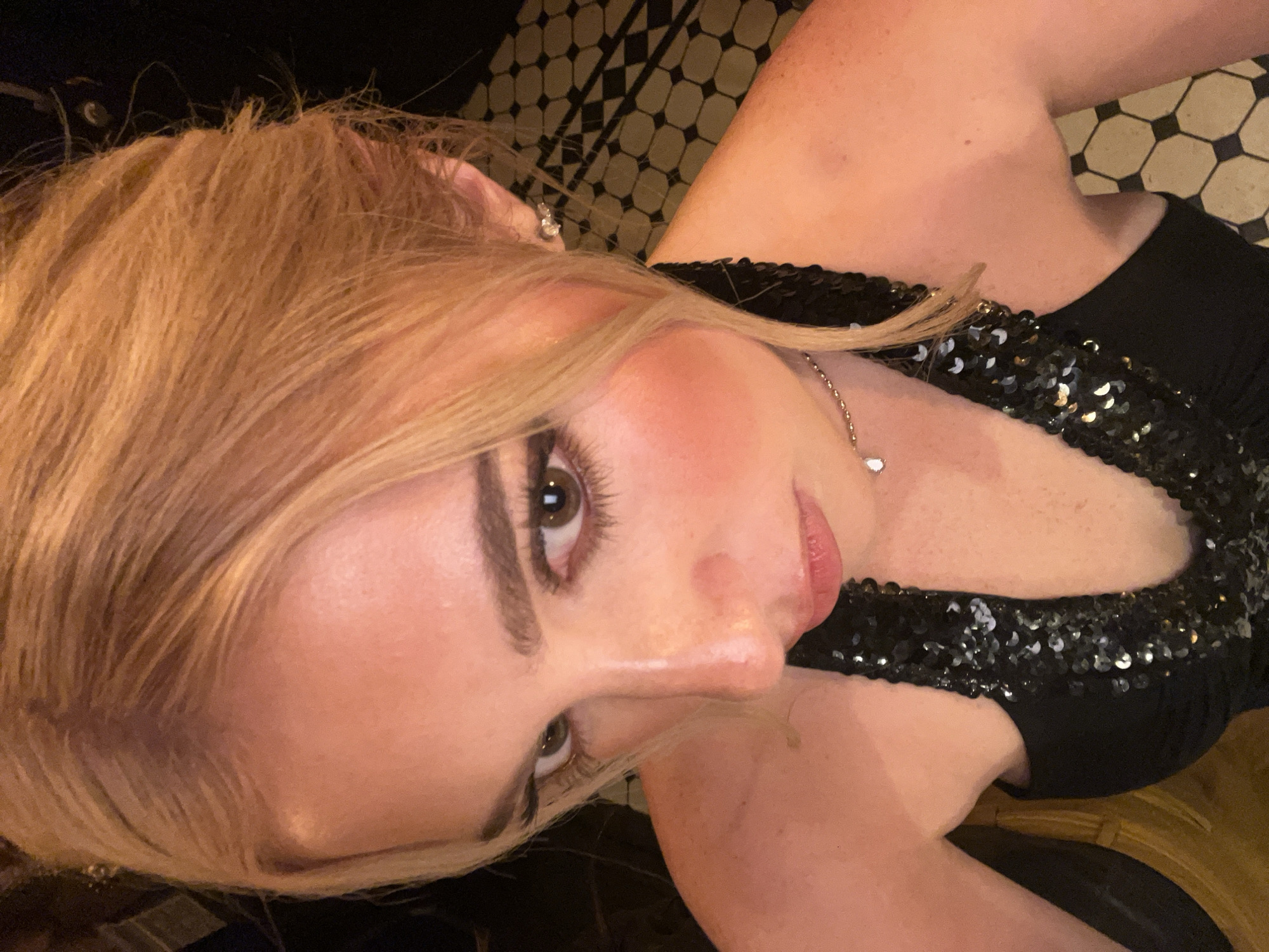 A person with light brown hair and fair skin poses in a selfie. They are wearing a black, sequined top and a delicate silver necklace. The person's face is partially turned, with one eye looking directly at the camera. The background features a patterned tile floor of white, black, and gold hues. The location is not possible to determine with available information.