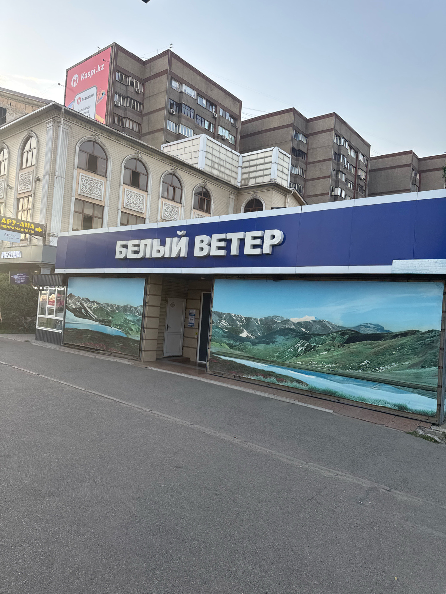 The image captures the facade of a retail establishment in Almaty, Kazakhstan. The business, identified by the Cyrillic lettering "БЕЛЫЙ ВЕТЕР" (White Wind), is prominently displayed on a large blue awning. Below the awning, the storefront is covered by large murals depicting a scenic mountain landscape with a river. To the left of the main storefront, there is a smaller kiosk with windows, and further left, the building's architecture is visible, featuring arched windows with decorative elements and what appears to be signage for "Kaspi.kz," a Kazakhstani fintech company, on an upper level. The street in front of the building is paved, and a sidewalk is present. The surrounding buildings are multi-story structures, typical of an urban environment. The sky is overcast, suggesting an outdoor scene during daylight hours.