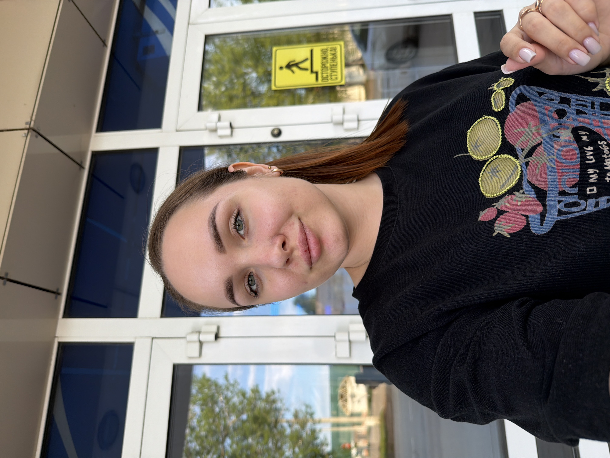 A woman is in front of a building. The yellow sign on the door has the words, "ОСТОРОЖНО! СТУПЕНЬКА!" on it with an illustration of a person descending stairs. A street is reflected in the windows of the building.