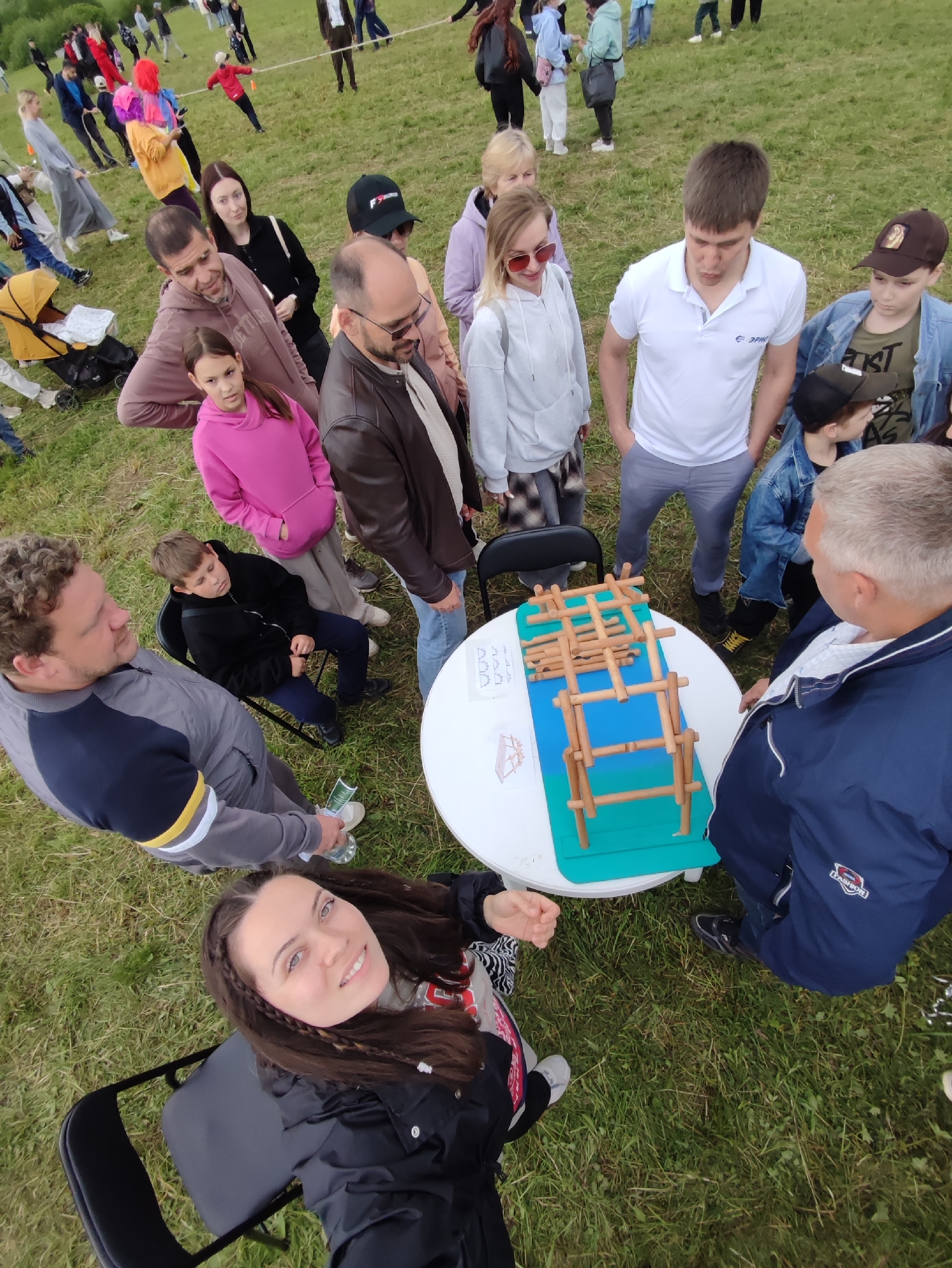 The image depicts an outdoor gathering on a grassy field in Unknown, Unknown. Multiple individuals are arranged around a white circular table, which displays a complex wooden construction resembling a bridge or tower, set upon a blue and green mat, alongside a piece of paper with diagrams.
On the far left, a man with curly brown hair and a beard, wearing a grey and navy striped top, holds a clear plastic bottle. To his right, a young boy in a black hoodie and dark pants sits on a black folding chair, looking down. Adjacent to him, a man with short dark hair and a beard wears a brown leather jacket over a light shirt, observing the table. A young girl in a bright pink hoodie stands next to him, also looking at the table. Behind them, a woman with dark hair in a black long-sleeve top looks towards the table. Further right, a woman with blonde hair, wearing a grey hoodie and sunglasses, stands near a man in a white polo shirt with an "ЭРНЕ" logo and grey pants, both focused on the table. Next to them, a boy in a denim jacket and brown baseball cap, and another child in a black baseball cap, also look down. On the right side of the table, an older man with grey hair, dressed in a navy blue jacket with white stripes, observes the wooden structure.
In the immediate foreground, a young woman with long, braided dark hair, wearing a black jacket over a patterned shirt, sits on a black folding chair, looking directly at the camera with a slight smile.
In the background, numerous people are scattered across the grassy field, some engaged in what appears to be a tug-of-war game, including a person in a red outfit pulling a rope and another in a multi-colored outfit. A yellow baby stroller is visible in the mid-ground on the far left. The ground is primarily green grass with some sparse patches. The scene is illuminated by natural light, indicating daytime.