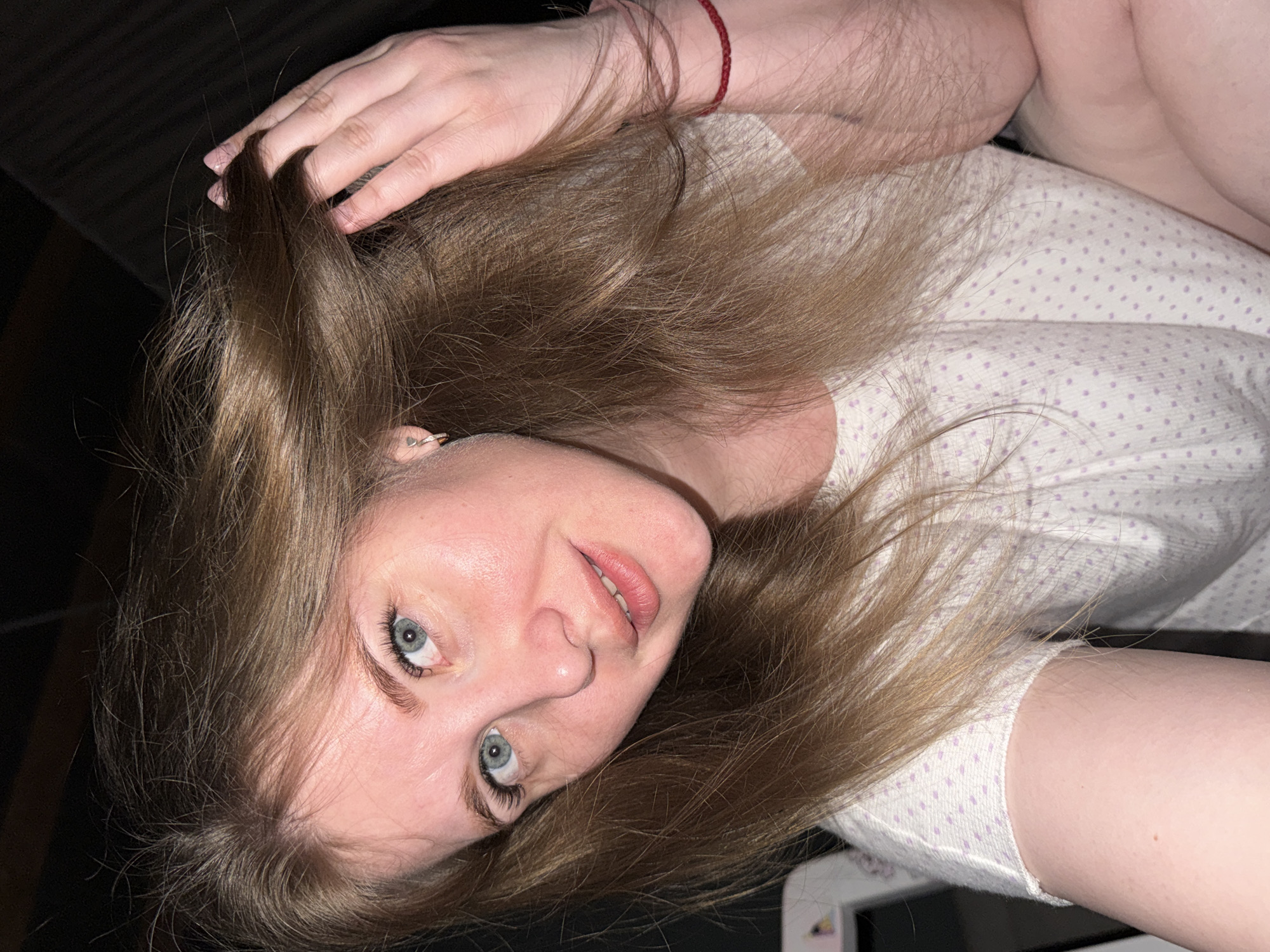 The image shows a person with fair skin, light brown hair, and blue eyes. They are wearing a white shirt with purple dots. A red string bracelet is visible on their wrist. The background is dark and indistinct. The person is looking at the camera.