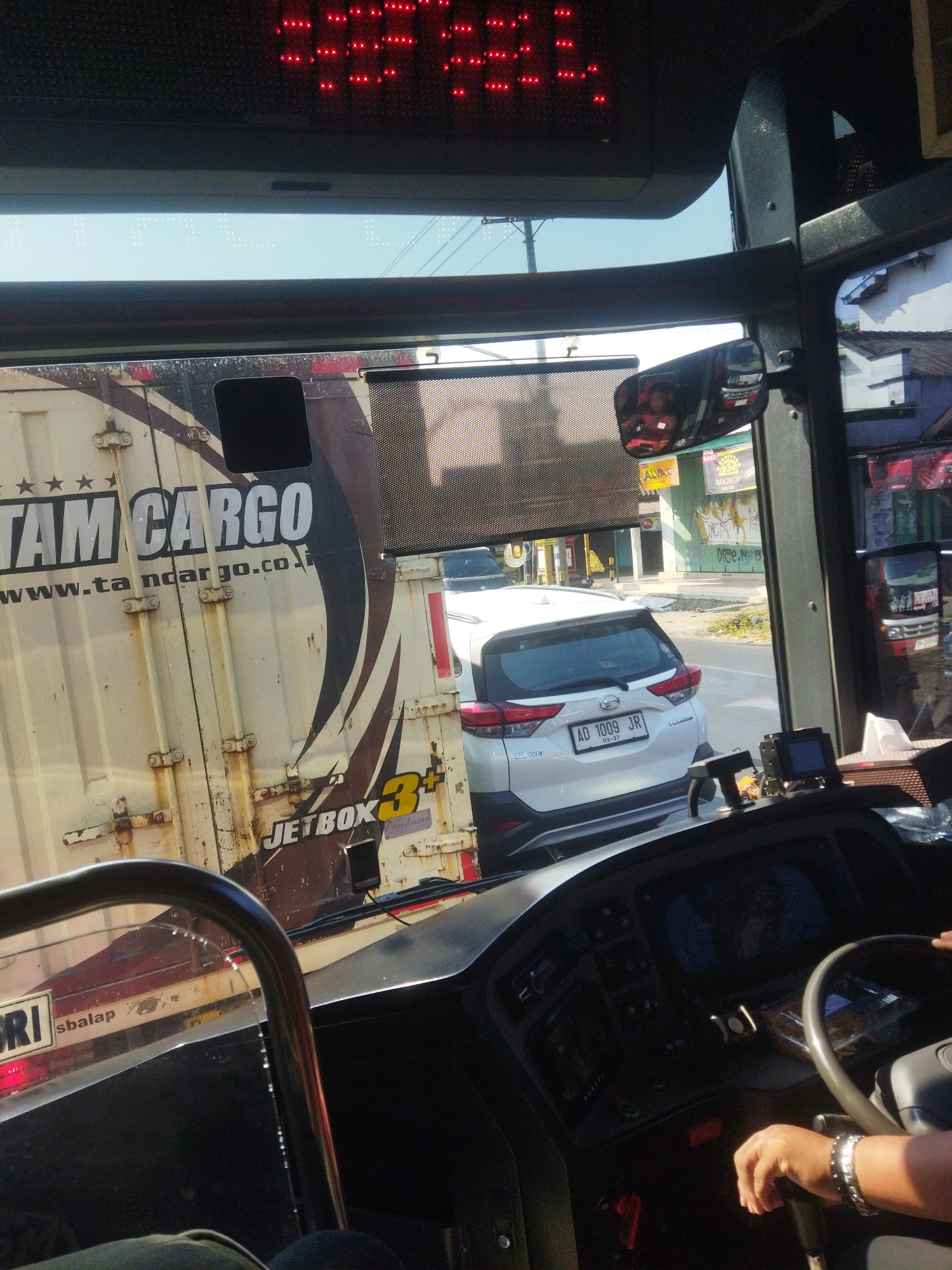 The image shows the interior of a bus, viewed from the driver's seat. The driver's hands are on the steering wheel and gear shift. Outside the bus, a white SUV with license plate AD 1009 JR is visible, as well as a shipping container with "TAM CARGO" written on it. The scene appears to be on a sunny day, with buildings and trees visible in the background. The location is identified as Begajah, Indonesia, though no specific visual cues in the image directly indicate this location. The overall impression is that of a typical street scene in Indonesia, possibly during a bus journey.