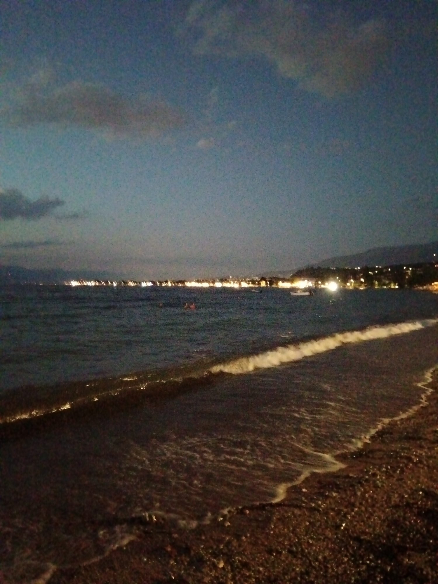 The image depicts a nighttime view of a beach along the Municipal Unit of Vrachneika, Greece. The sky is dark blue with scattered, soft clouds. The sea is calm with small waves gently lapping onto a pebble beach. In the distance, a coastal town is illuminated by numerous lights, creating a blurred line of brightness against the darker silhouette of hills or mountains. A few faint lights are visible on the water, possibly from boats. The overall scene is tranquil and atmospheric, characterized by the soft glow of the distant town and the subtle interplay of light on the water and shore.