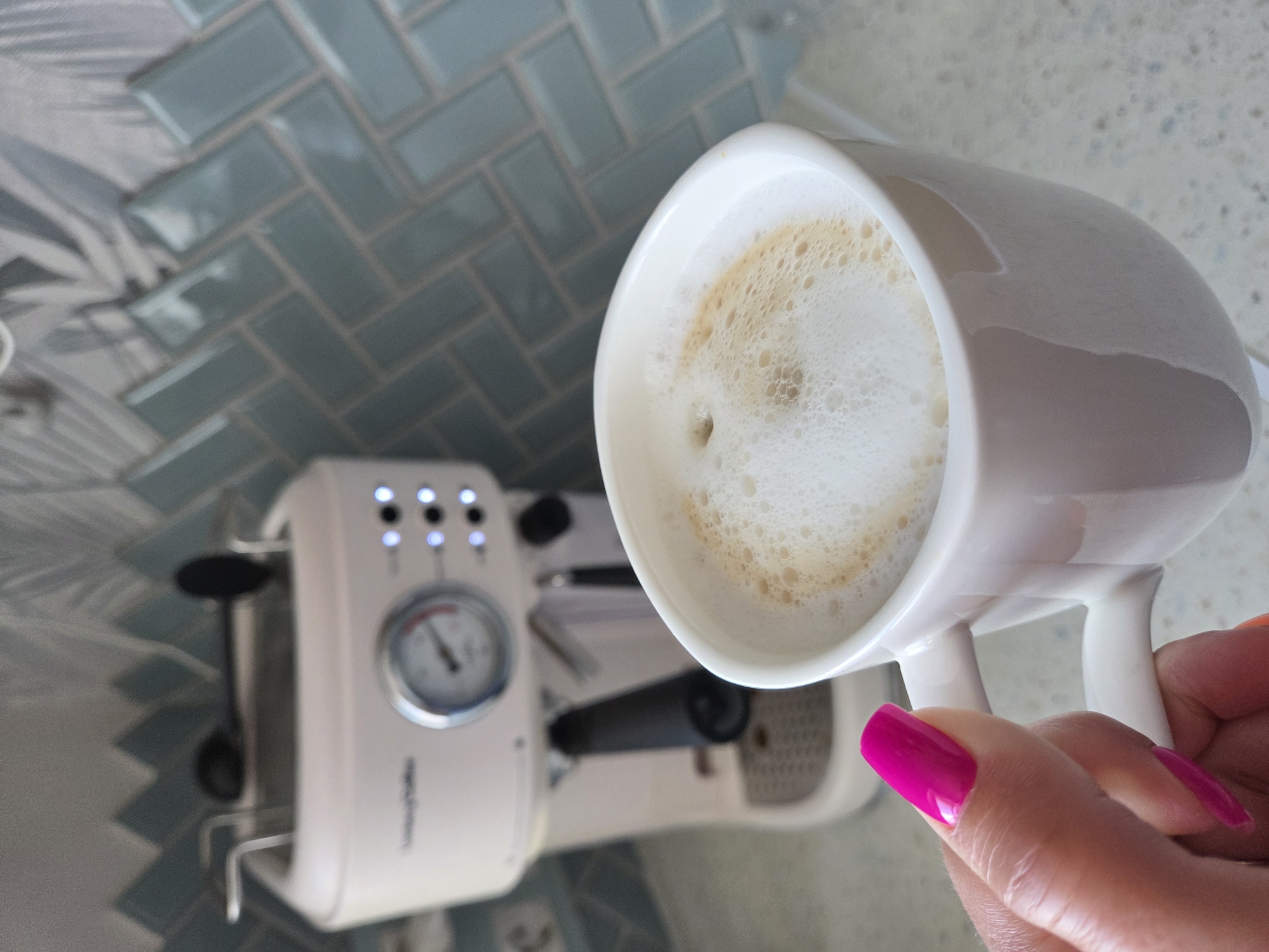 A close-up shot shows a white mug filled with a beverage topped with foam, held by a hand with bright pink nail polish. In the background, out of focus, is a cream-colored espresso machine with a pressure gauge and buttons. Behind the machine is a wall with bluish-gray, rectangular tiles laid in a herringbone pattern. The surface beneath the mug and espresso machine appears to be a light, speckled countertop. The location in the city of Birobidzhan, Russia, is not discernible from the image.