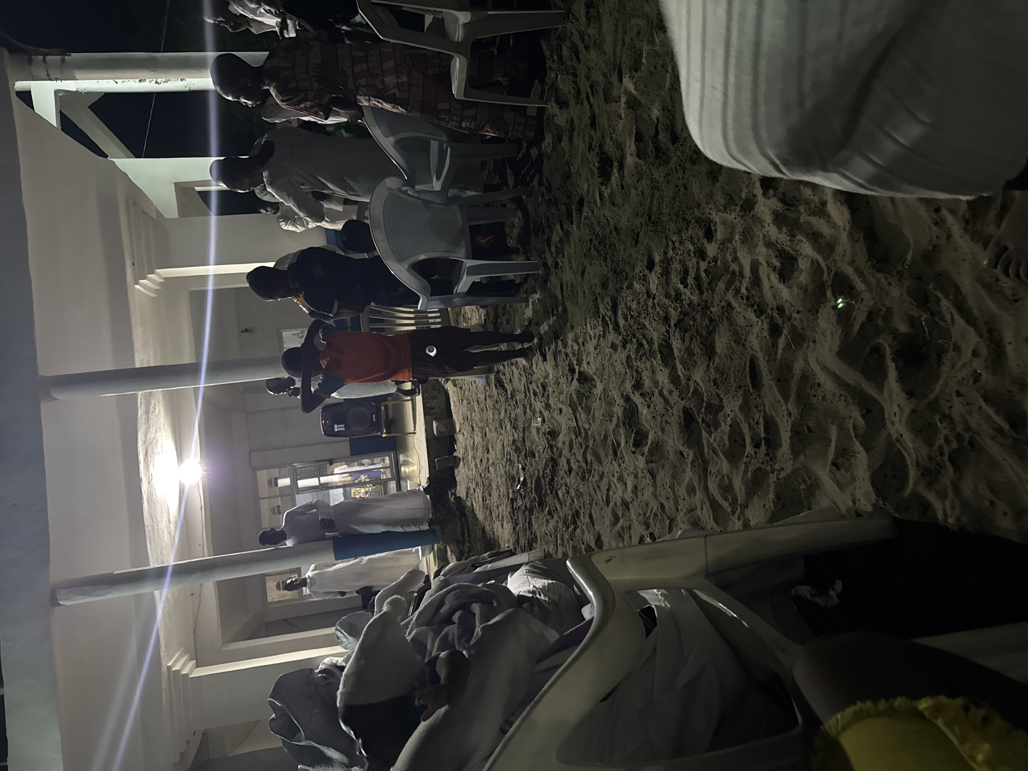 A low-angle, horizontally oriented photograph captures a gathering of people on a sandy ground adjacent to a white building with illuminated interiors and support pillars, likely at night. Individuals are standing and sitting on white plastic chairs. A person in an orange shirt is standing. Some people are wearing white clothing, while one person is wearing a patterned shirt. The location is suggested to be Badagry, Nigeria.