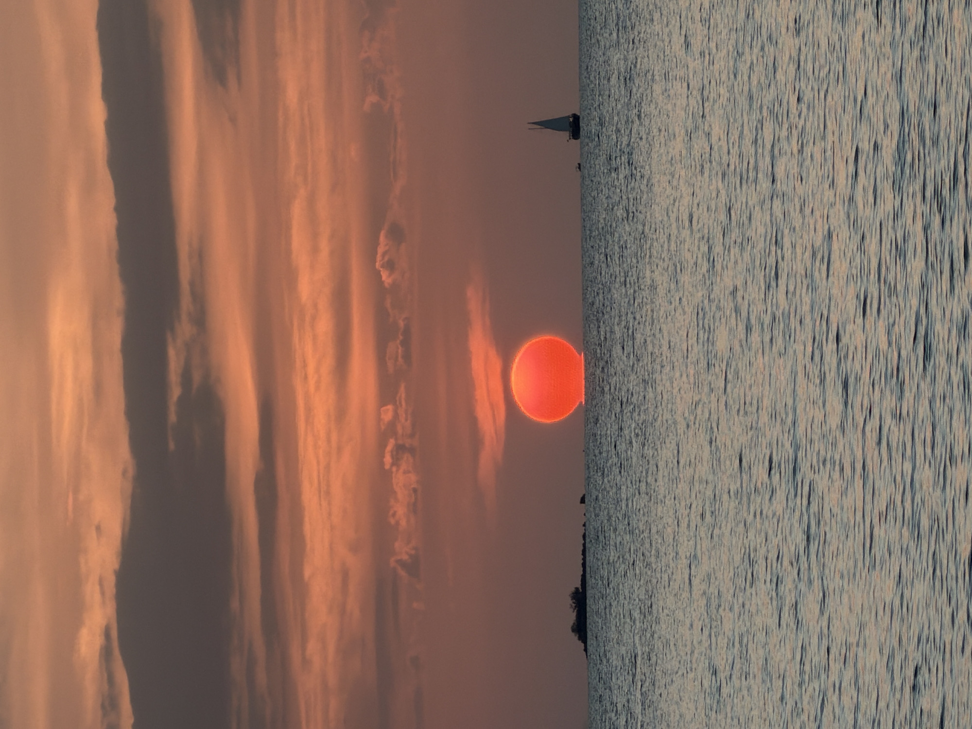 A wide shot captures a vibrant sunset over the Gulf of Thailand. The sun, a brilliant orange orb, is partially submerged at the horizon, casting a warm glow across the sky. The sky is filled with streaks of orange, pink, and gray clouds, creating a dramatic backdrop. In the distance, a silhouette of a small island with vegetation is visible on the left, and a sailboat with its sail raised glides across the water on the right. The sea is calm, reflecting the colors of the sky with gentle ripples. The scene appears to be captured from a beach or boat in the Ko Samui District of Thailand.