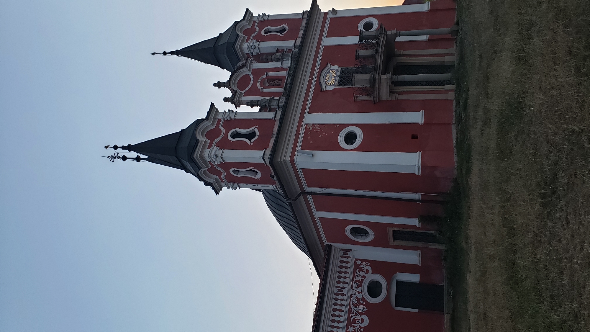 The image shows a red and white church with two prominent spires, a dome, and a balcony. The church is situated in a grassy field under a clear, pale sky, suggesting either early morning or late evening. The architectural style appears to be Baroque or Neo-Baroque, with decorative elements on the facade. The building seems aged, with some wear visible on the paint. There are no people visible in the image. The church is located in the city of Prešov, Slovakia. Specifically, it is the Church of the Exaltation of the Holy Cross, a Roman Catholic church in the town of Pušovce, which is part of the Prešov region.