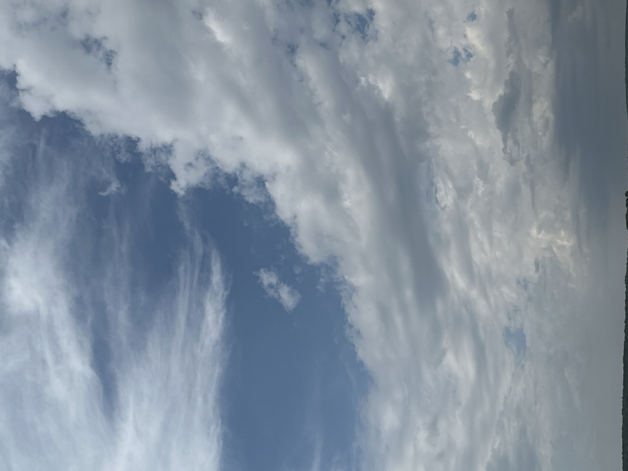 The image displays a wide expanse of sky over a low-lying landscape, likely the city of Makiv, Ukraine. The sky is filled with a variety of clouds, ranging from wispy cirrus formations in the upper left to dense, grey cumulus clouds dominating the right side and lower portions of the frame. Patches of clear blue sky are visible between the cloud formations. The horizon line, visible at the very bottom, suggests a flat terrain with some scattered vegetation or structures, but these are indistinct due to the distance and the focus on the sky. There are no people or discernible objects in the scene, and the overall impression is one of a natural, open environment.