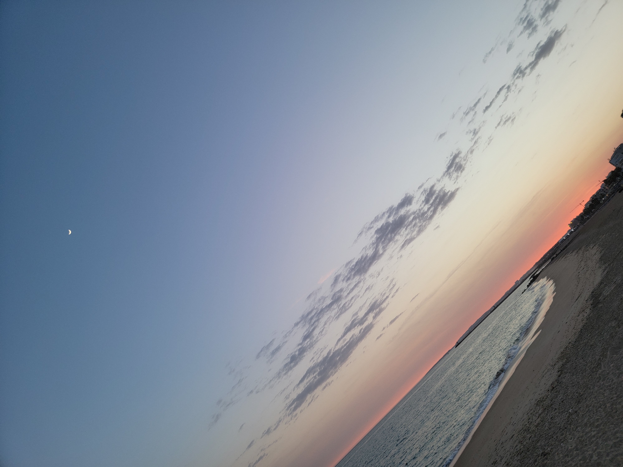 The image shows a diagonal view of a beach and the ocean at sunset in Quarteira, Portugal. The sky is a gradient of pale blue to soft pink and orange, with scattered clouds. A crescent moon is visible in the upper right portion of the sky. The ocean on the left is a deep turquoise color, with gentle waves lapping at the sandy shore. The beach is a muted brown color, and the shoreline extends into the distance where a town or city is visible along the coast, with lights starting to appear. There are no people or specific objects prominently featured. The overall atmosphere is tranquil and serene, characteristic of a coastal sunset.