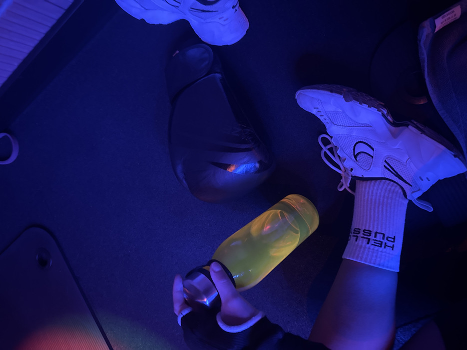 Under blue lighting, a workout scene unfolds. A white sneaker rests on a foot clad in a white sock with black lettering, near a yellow liquid-filled bottle held by a hand in a black glove with white trim. Adjacent to these are a pair of black boxing gloves and another white sneaker, positioned on a dark surface that includes a portion of a yoga mat.