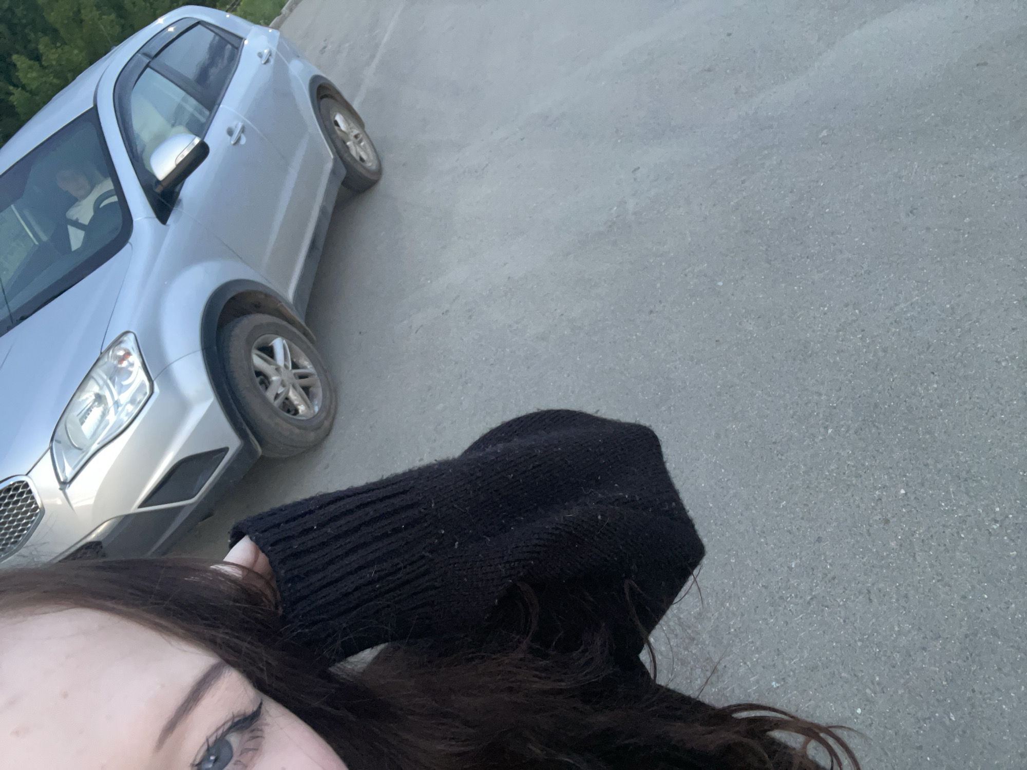 The image presents a first-person perspective, dominated by a paved ground and featuring a partial view of a person and a silver SUV. In the bottom left corner, the left side of a person's face is visible, showing a light-colored eye, eyebrow, and cheek, framed by long, dark hair. A black, ribbed sweater sleeve extends into the right side of the frame, notably displaying several small white specks, possibly lint or dust.
A silver or light grey SUV is parked on the left side of the frame, angled slightly towards the upper right. Its front-left headlight, part of the grille, and the side profile with dark window deflectors are clearly visible. A blurry figure can be discerned inside the driver's side of the vehicle. The SUV's wheels have multi-spoke rims.
The ground surface is a light grey asphalt or concrete, appearing flat and consistent. In the distant background, behind the vehicle, hints of green foliage suggest the presence of trees or bushes. The lighting indicates it is daytime, possibly late afternoon or early evening, with soft, even illumination and an absence of harsh shadows. There is no discernible text within the image. The scene is an outdoor, likely urban or suburban, setting in Votkinsk, Russia.