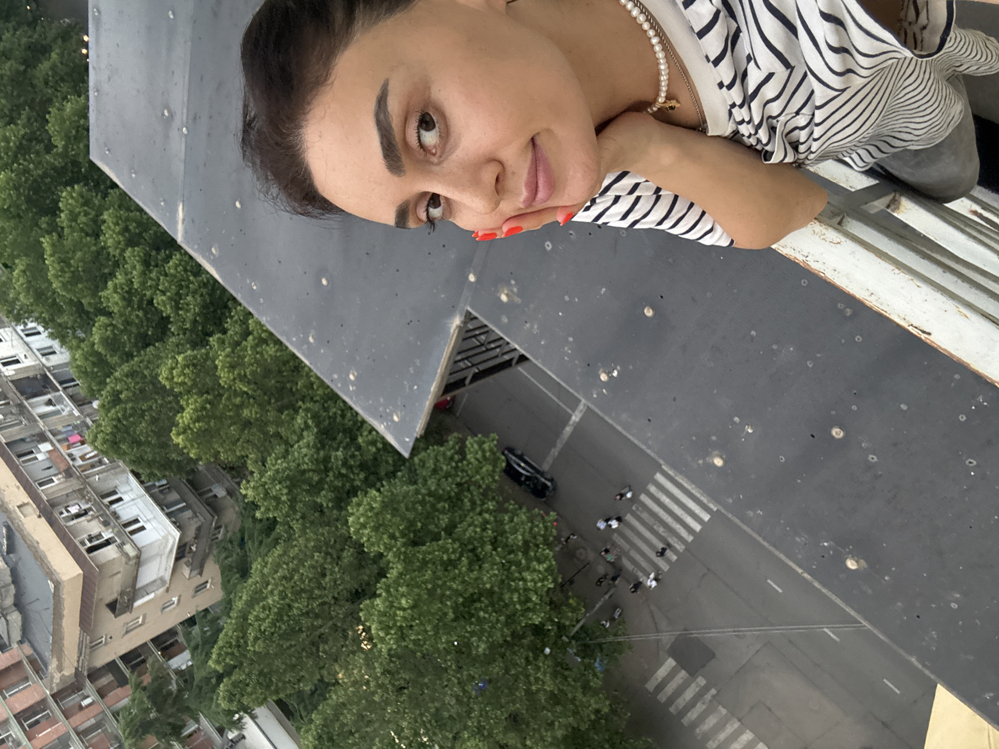 A medium shot, taken from a high vantage point, shows a woman with dark hair and brown eyes posing for a selfie. She is leaning her head on her hand, and her fingernails are painted red. She is wearing a striped t-shirt and a pearl necklace. In the background, there are trees and buildings, and a street with people and cars. The image appears to have been taken in an urban environment, possibly Tbilisi, Georgia, given the architecture and vegetation.