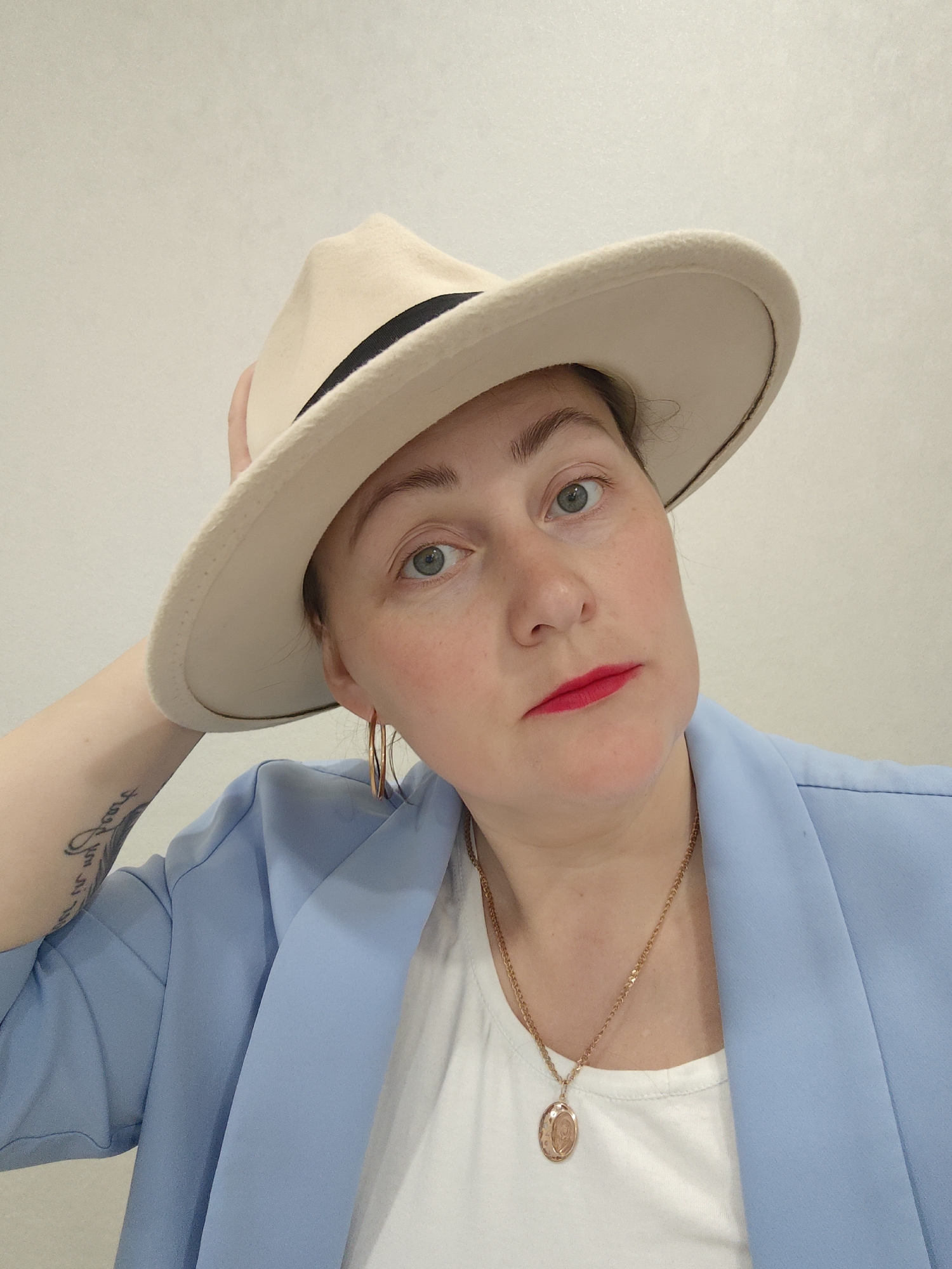 A medium-shot portrait features a woman with light skin, light blue/grey eyes, and a neutral, direct expression. She is wearing bright red lipstick and some blush. She is dressed in a light blue blazer over a white t-shirt. On her head is a light beige fedora-style hat with a black band, which she holds with her left hand, her fingers touching the back of the hat. She is accessorizing with gold-colored hoop earrings and a gold chain necklace with an oval locket-style pendant. A tattoo of decorative script is visible on her left forearm. The background is a plain, light off-white wall, indicating an indoor setting with bright, even lighting. The scene is located in Markovskii, Russia.
