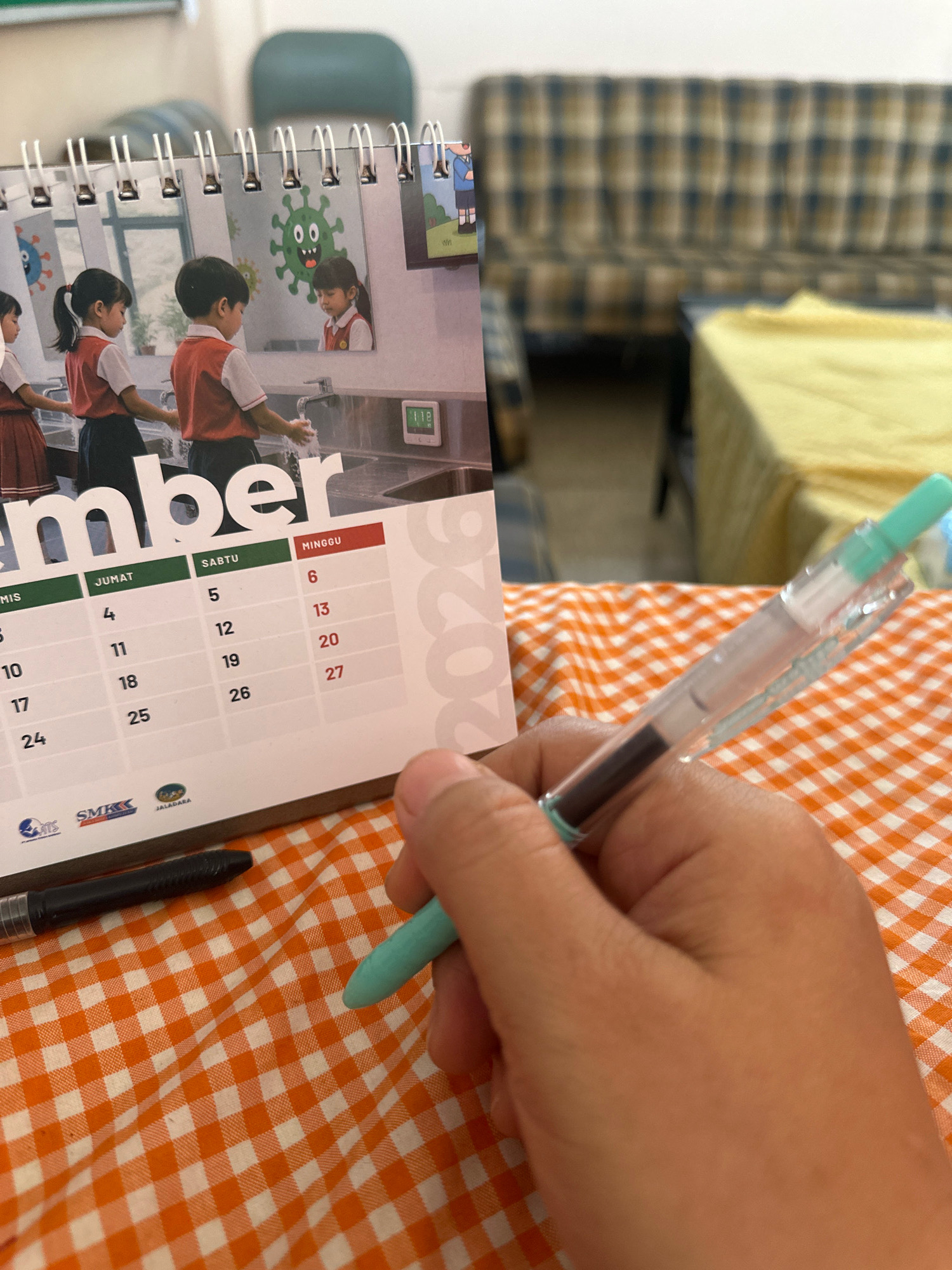 The image depicts a close-up view of a hand holding a turquoise pen, poised over a calendar that is partially visible. The calendar appears to be for the month of December, as indicated by the large "mber" text on the left side of the frame. The calendar itself shows a grid with days of the week labeled in Indonesian: "JUMAT" (Friday), "SABTU" (Saturday), and "MINGGU" (Sunday). The dates visible include the 4th, 5th, 6th, 10th, 11th, 12th, 13th, 17th, 18th, 19th, 20th, 24th, 25th, 26th, and 27th. Below the calendar dates, there are logos for "ATS," "SMKK," and "JALADARA." The calendar also features an illustration of children in school uniforms washing their hands, with a cartoon virus image in the background. The background of the scene includes a couch with a plaid pattern and a yellow-covered table. The surface on which the calendar and pen rest is a tablecloth with an orange and white checkered pattern. The image is taken indoors. No specific location in the city of Purwotengah, Indonesia can be identified from the visual information provided.