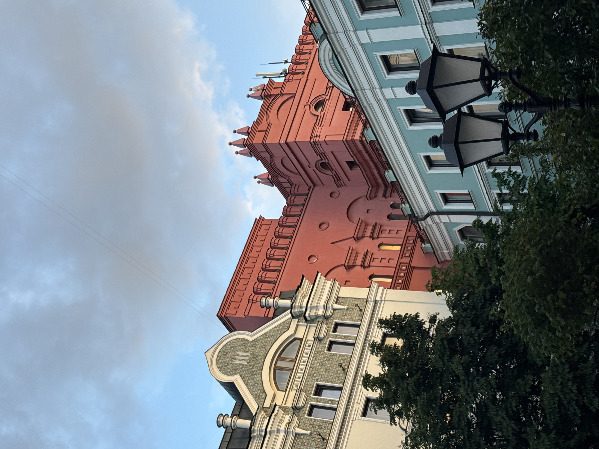 A low-angle shot depicts three buildings in Moscow, Russia. The building in the center is red with distinct architectural features. The building to the right is a lighter shade of blue with window frames. A portion of the building to the left is visible, featuring a curved top and tiles. Street lamps and tree foliage are in the foreground. The sky is cloudy.