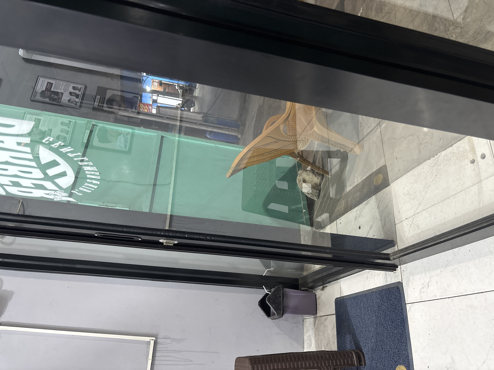 The image depicts the exterior of a barbershop, with its entrance featuring a black frame and glass doors. Through the glass, a light brown woven plastic chair is visible inside, positioned on a tiled floor. A large, light green wall is also seen, adorned with posters displaying hairstyles and men. To the left of the door, on the exterior, a dark brown woven plastic ottoman and a purple trash can are present. The floor outside the shop is tiled with white and grey marble-like patterns. The scene is located in Wanajaya, Indonesia.