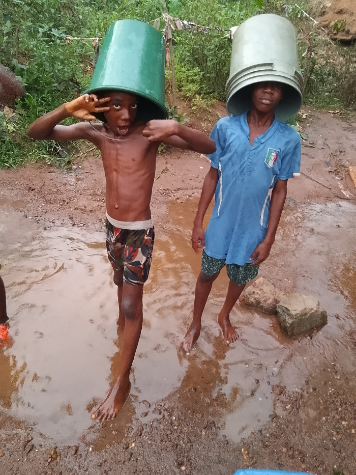 Two young boys stand in a muddy puddle. Each has a plastic bucket on their head. The boy on the left is shirtless, wearing patterned shorts, and making a gesture with his hands. The boy on the right wears a blue t-shirt and green shorts. Their feet are submerged in the murky water. The background features greenery and soil, suggesting an outdoor, possibly rural, setting. The scene is playful and candid.
