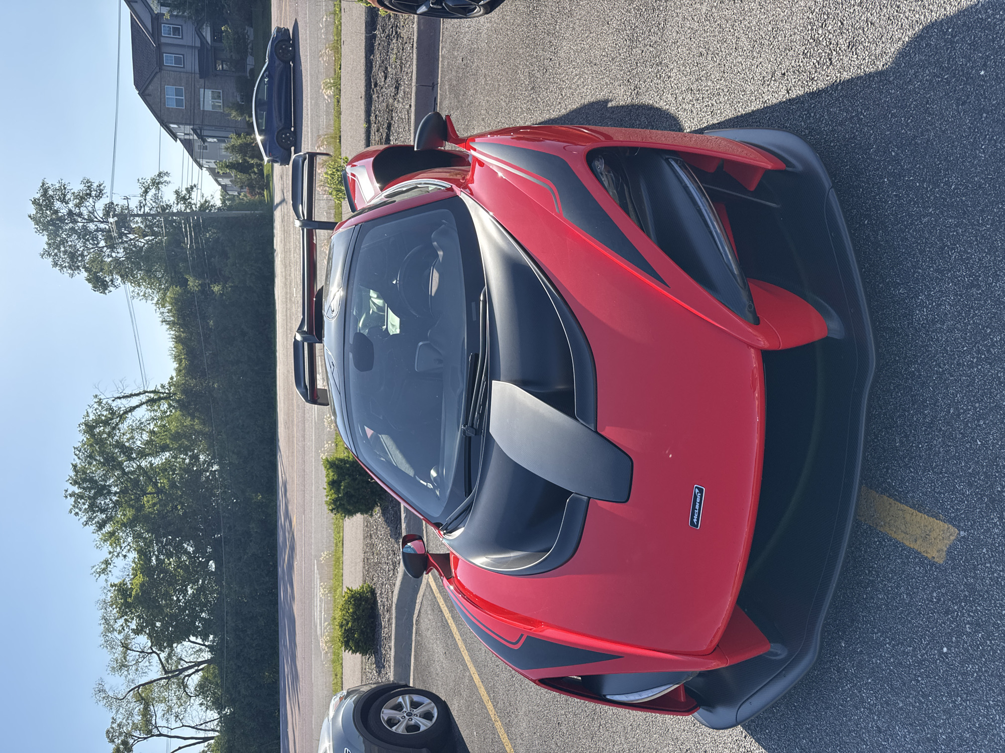 The image depicts a red McLaren sports car parked in a parking lot. The car has black accents and a prominent rear wing. There are other vehicles parked nearby. The location appears to be Warrenville, United States of America. A building and trees are visible in the background, suggesting an urban or suburban setting. The sky is clear.