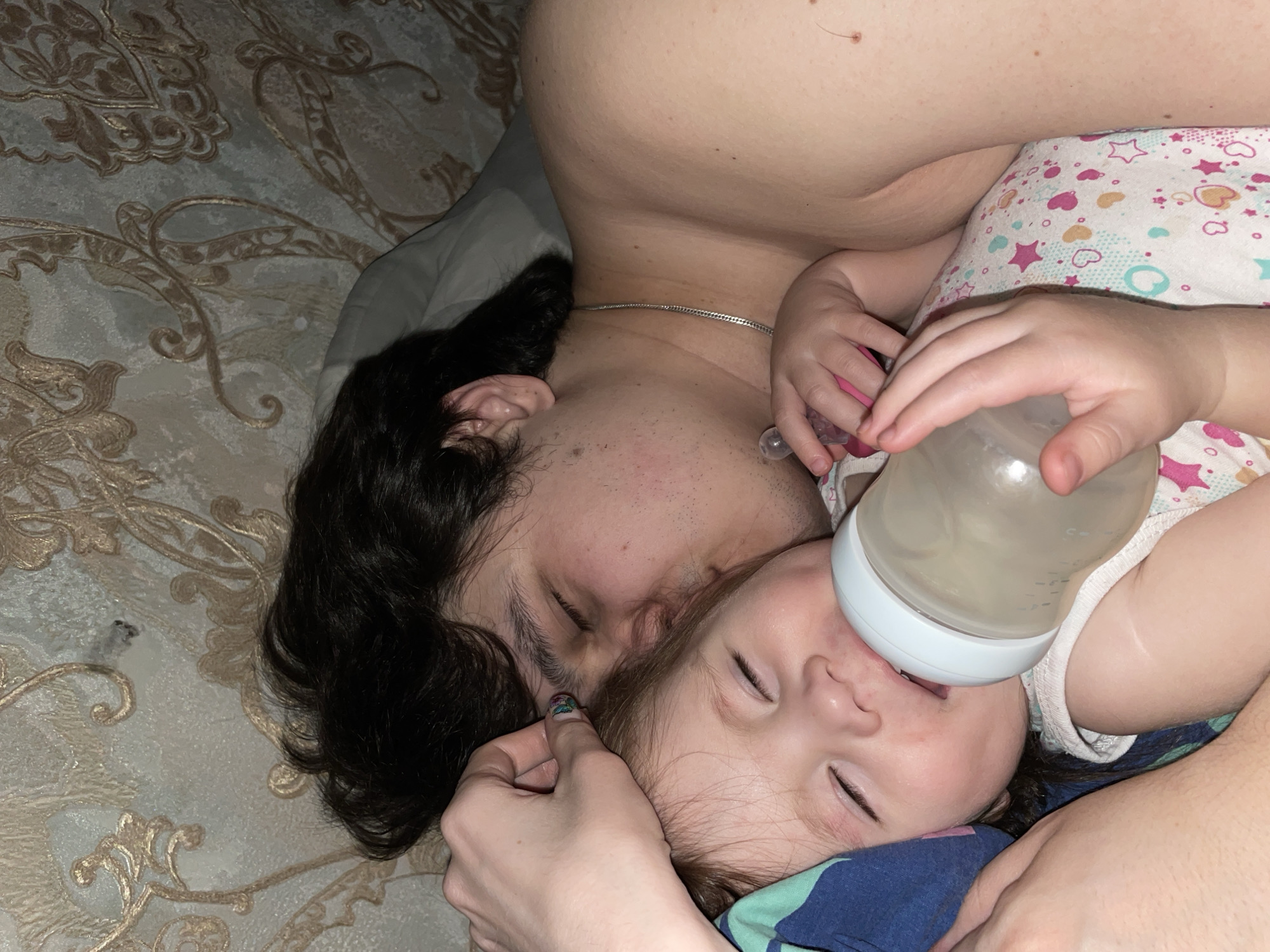 A man and a baby are lying on a bed. The man has dark hair and is wearing a chain. The baby is wearing a patterned outfit and drinking from a bottle. A hand is holding the baby’s head. The location is not identified as being in Izhevsk, Russia.