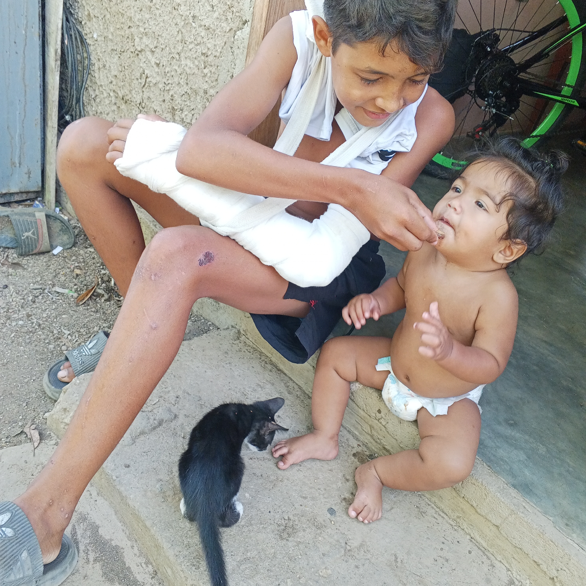 An outdoor, close-up shot shows two young children and a black and white kitten. On the left, a boy with dark hair and brown eyes, wearing a white tank top, is seated. His right arm is in a cast and wrapped with white bandages, and his left arm is also bandaged. His left knee has a scraped wound with dried blood. A kitten is curled up on the concrete in front of the boy. To the boy's right, a baby is seated and wearing a diaper. The boy is holding a small piece of food to the baby's mouth. A bicycle with green accents is visible in the background. The scene appears to be in a residential outdoor area with a textured wall and concrete ground.