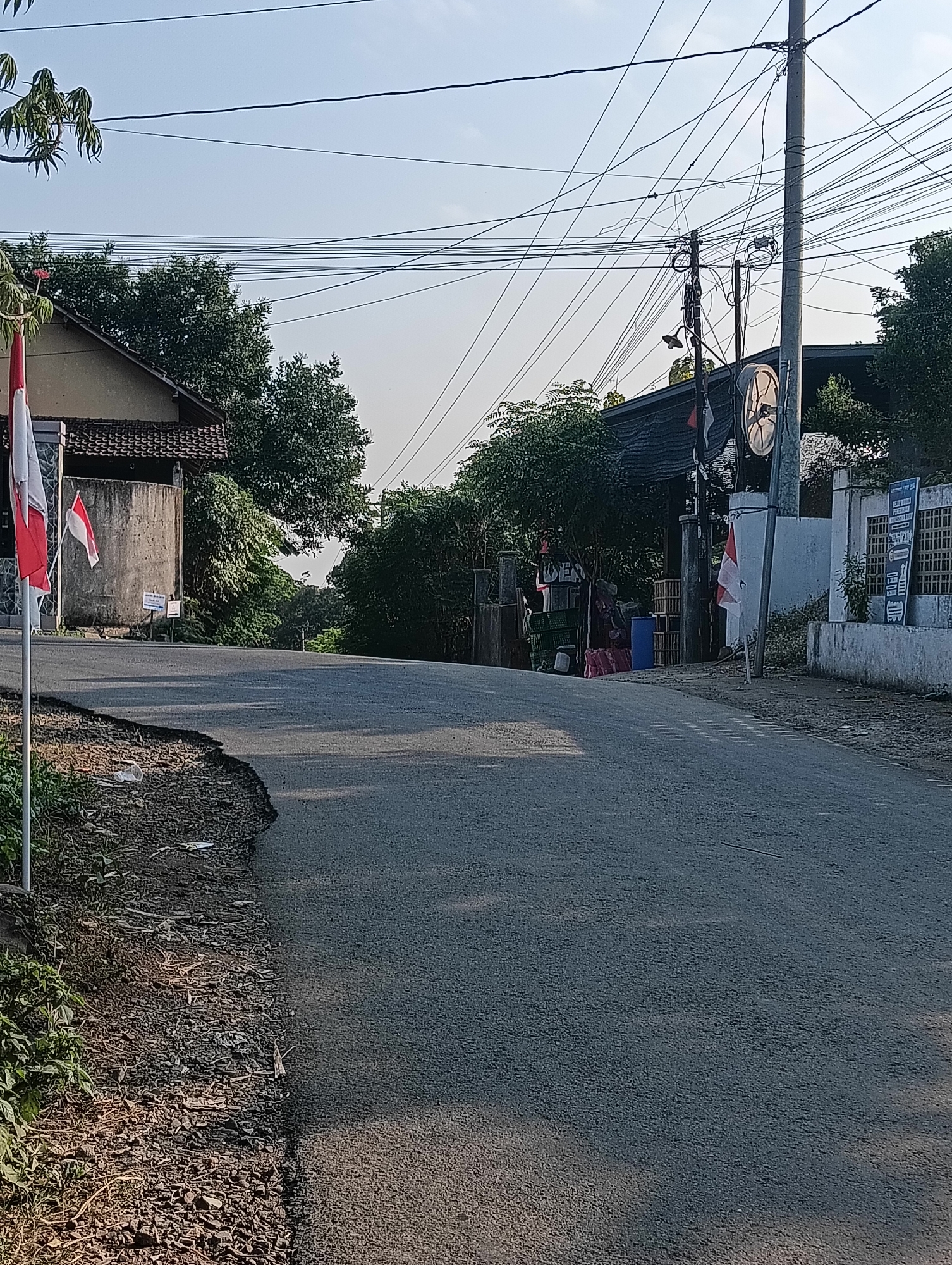 The image depicts a paved road curving to the right, bordered by a gravel and grassy shoulder on the left and a concrete wall with a fence on the right. Two Indonesian flags are planted on the left shoulder. Several trees and buildings are visible in the background, along with numerous power lines and a utility pole. The sky is clear and blue, suggesting daytime. The scene appears to be in a semi-rural or residential area in Cluwak, Indonesia. There are no people visible in the image, and no specific actions or interactions are taking place. Objects present include flags, a road, utility pole, power lines, trees, buildings, a concrete wall, and a fence. The overall environment is a street scene under a clear sky.