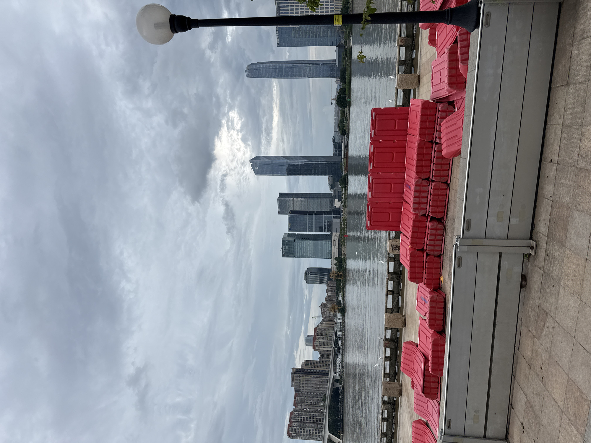 The image depicts a waterfront scene in Tianhe District, China, under a cloudy sky. In the foreground, a series of modular flood barriers, likely constructed from grey metal panels, are installed along a paved walkway. Behind these barriers, a large quantity of bright red plastic barriers are stacked in neat piles. A river runs between the foreground and the opposite bank, where a city skyline is visible. The skyline is dominated by numerous modern skyscrapers of varying heights and architectural styles. A bridge extends across the river in the mid-ground, connecting to the cityscape. Several white birds are perched on a low stone wall bordering the river. A lamppost with a globe-shaped light fixture stands on the right side of the image, its dark pole contrasting with the muted tones of the sky and water. The overall impression is one of an urban waterfront, possibly prepared for potential flooding or undergoing construction, with a backdrop of contemporary city architecture.