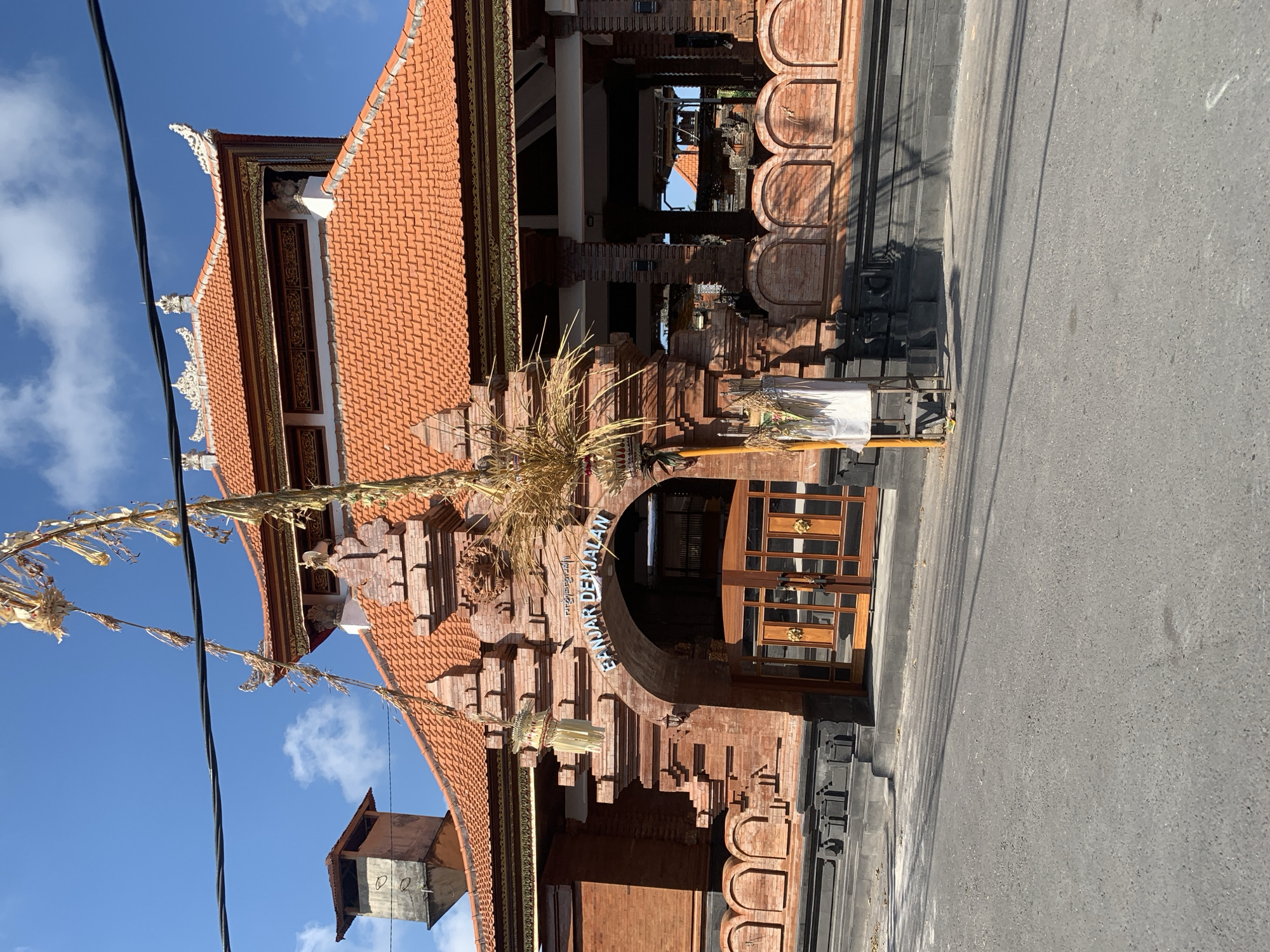 An exterior, eye-level, medium shot captures the entrance to a building in Sukawati, Indonesia. The building features traditional Balinese architecture with red tile roofing and red brick walls. An arched entryway with wooden double doors is visible. Above the arch, blue lettering spells out "BANJAR DENJALAN". Decorative religious offerings hang from a pole in front of the building. Power lines are visible in the upper portion of the frame, and a paved road runs in front of the structure. The sky is blue with scattered white clouds. The scene is well-lit by sunlight, casting shadows.