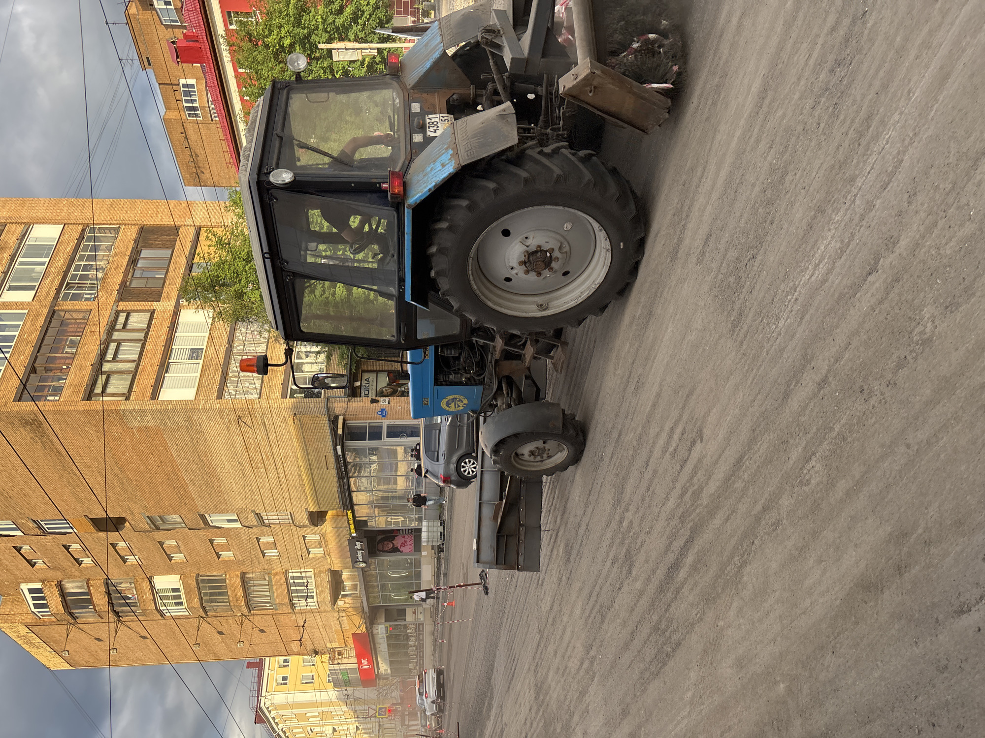 This image depicts a street cleaning operation in Murmansk, Russia. A blue tractor with a sweeping attachment is actively cleaning a dusty road. In the background, a multi-story brick building dominates the skyline, typical of Soviet-era architecture. Several cars are visible on the street, and pedestrians are walking along the sidewalk in front of shopfronts. The scene suggests an urban environment undergoing maintenance or preparation for road surfacing. The overall impression is one of everyday urban life with a focus on infrastructure work.