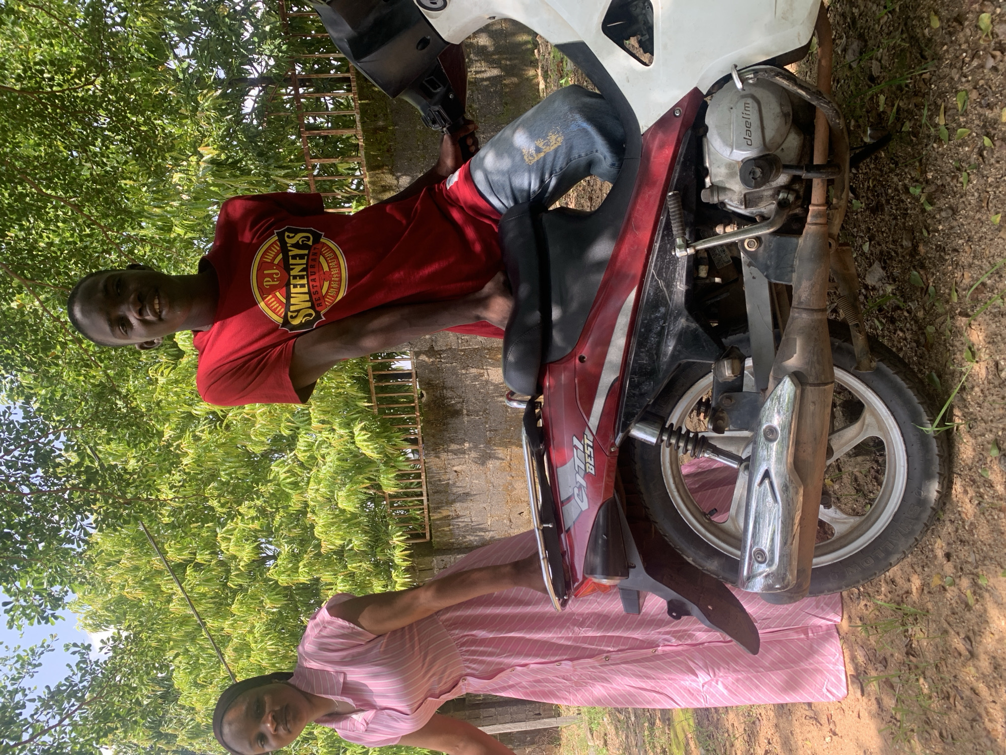 The image depicts two individuals, a young man and a young woman, outdoors in what appears to be a rural or semi-rural setting in Akwanga, Nigeria. The young man is seated on a motorcycle, leaning slightly to his left. He is wearing a red t-shirt with a circular logo featuring the text "P.J. SWEENEY'S RESTAURANT" in a stylized font. He is also wearing blue jeans. The young woman is standing beside the motorcycle, partially obscured by the vehicle and the foliage. She is wearing a pink and white striped dress.
The motorcycle is dark red and white, with visible engine components and a silver exhaust pipe. The ground around the motorcycle is covered with dirt, leaves, and some grass. Lush green trees with dense foliage are prominent in the background, suggesting a warm, sunny day. The lighting indicates it is daytime, likely midday or early afternoon. The overall mood appears casual and relaxed.
Visible text on the motorcycle includes "BEST" in white lettering on the side panel. The engine casing also shows the brand name "daelim". The scene is framed by greenery, with hints of a concrete wall and a metal fence visible in the background, further indicating an outdoor environment.