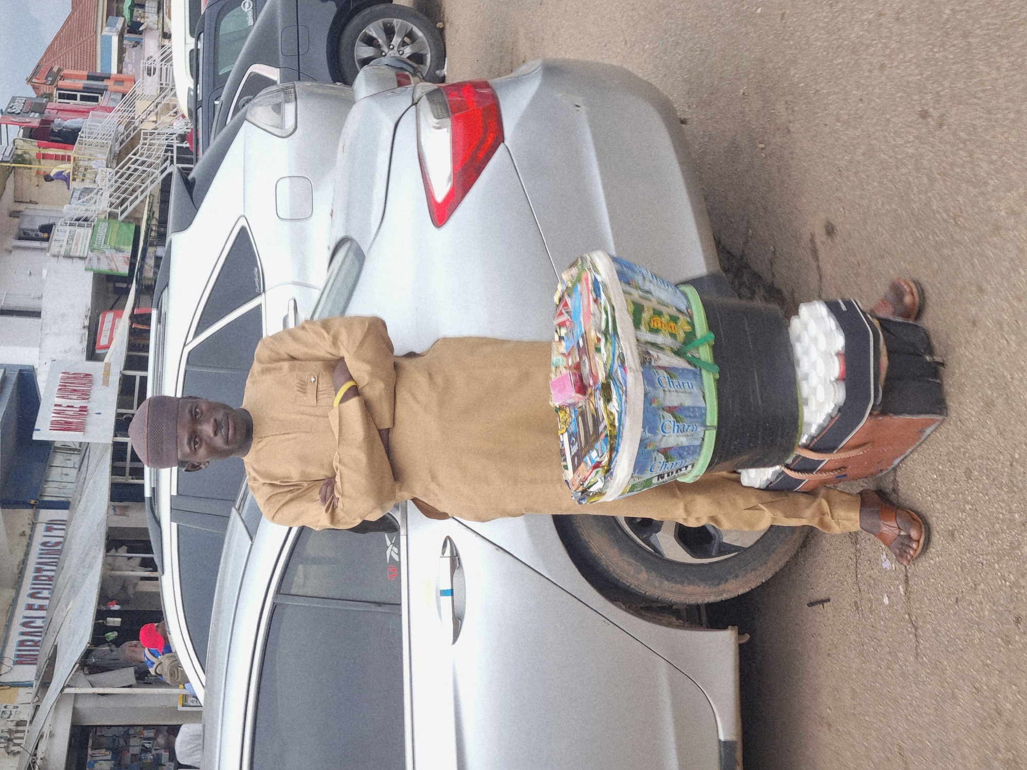 A man, appearing to be in his late 20s or early 30s, stands by a silver car. He is wearing a tan traditional outfit with a matching cap. His arms are crossed, and he is looking directly at the camera with a neutral expression. On his right side, he is carrying a large black bucket strapped to his back, which appears to be filled with various items, including what looks like packaged goods with colorful wrappers. A portion of the bucket's contents is visible, showing labels with text in a language that appears to be English or a similar Latin-script language. One prominent label reads "Charu."
The setting appears to be an urban environment in Jos, Nigeria. Behind the man and the car, there are buildings with signs, including one that reads "MIRACLE CURTAINS LTD." The buildings suggest a commercial area with shops. The ground is paved, and there are other vehicles visible in the background, including a black truck. The lighting suggests it is daytime, likely overcast or with diffused sunlight. The overall scene depicts a person engaged in some form of trade or transport, possibly a vendor or delivery person.