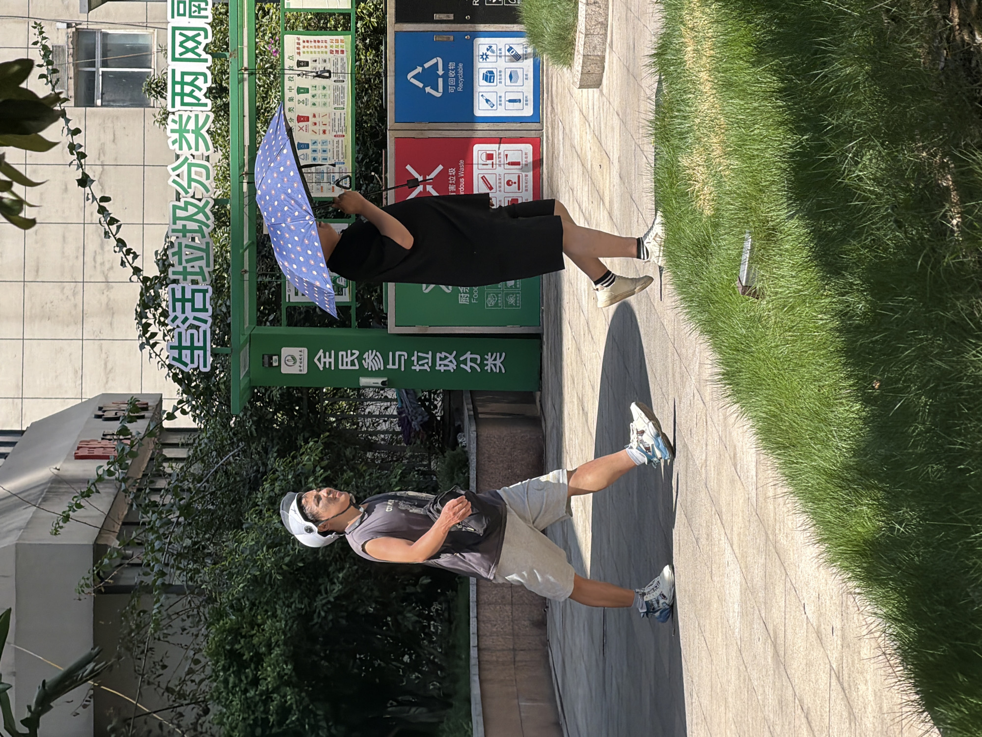 The image shows two people walking on a paved path in Yuzhong District, China. In the foreground, a person wearing a white helmet and a sleeveless grey t-shirt walks with a black bag slung across their chest, paired with grey shorts and athletic shoes. To their right, another person, dressed in a black outfit, walks while holding a blue polka-dotted umbrella. Behind them, a row of recycling bins is visible, including green, red, and blue bins, with signage indicating waste sorting in Chinese characters. A green structure with more signage stands above the bins. The environment includes lush green grass on the right side of the path and a building with trees and foliage in the background.